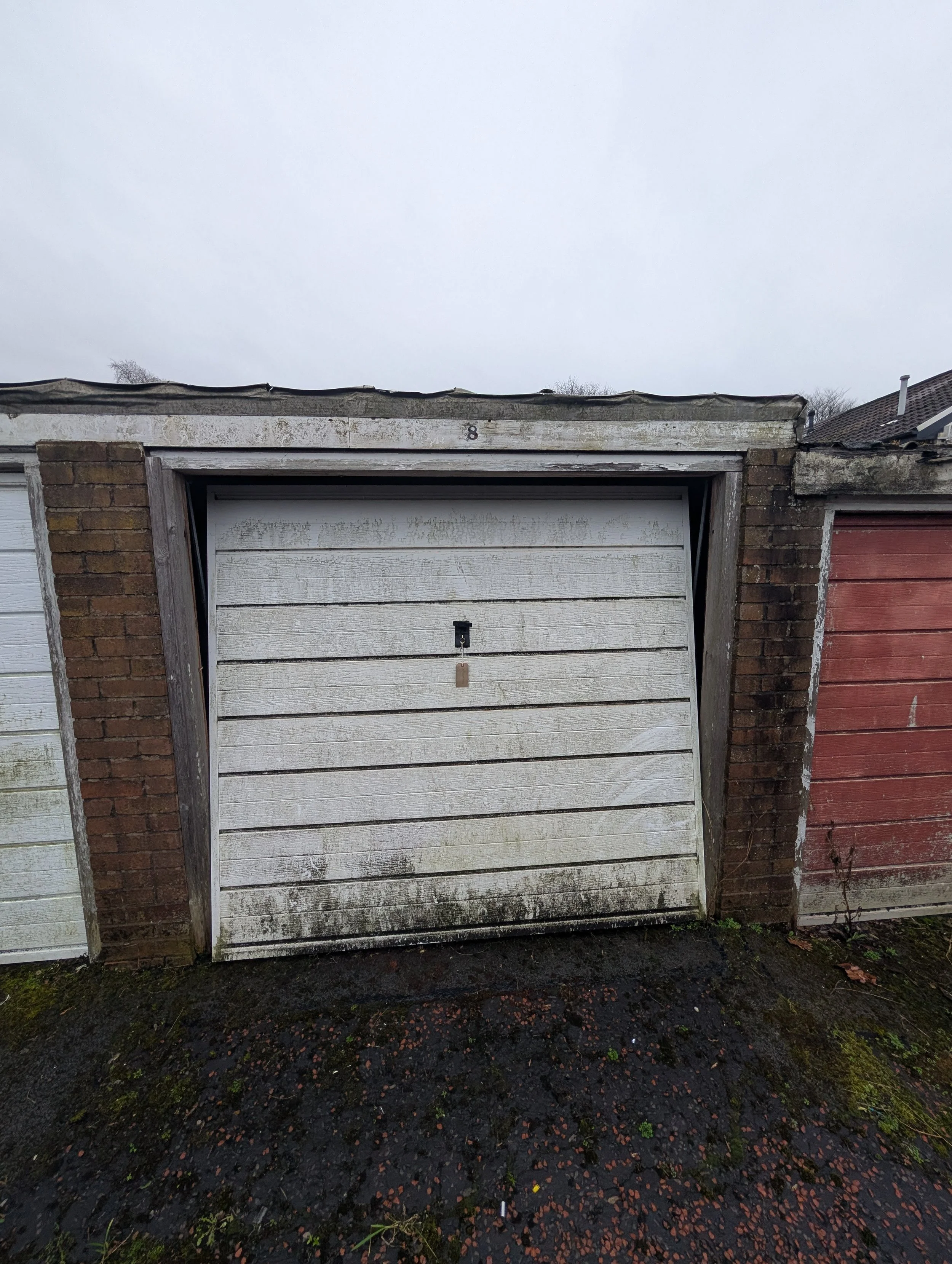 A weathered white garage door with dirt and grime at the bottom, flanked by brick walls, with another garage door on the right, all under a cloudy sky.
