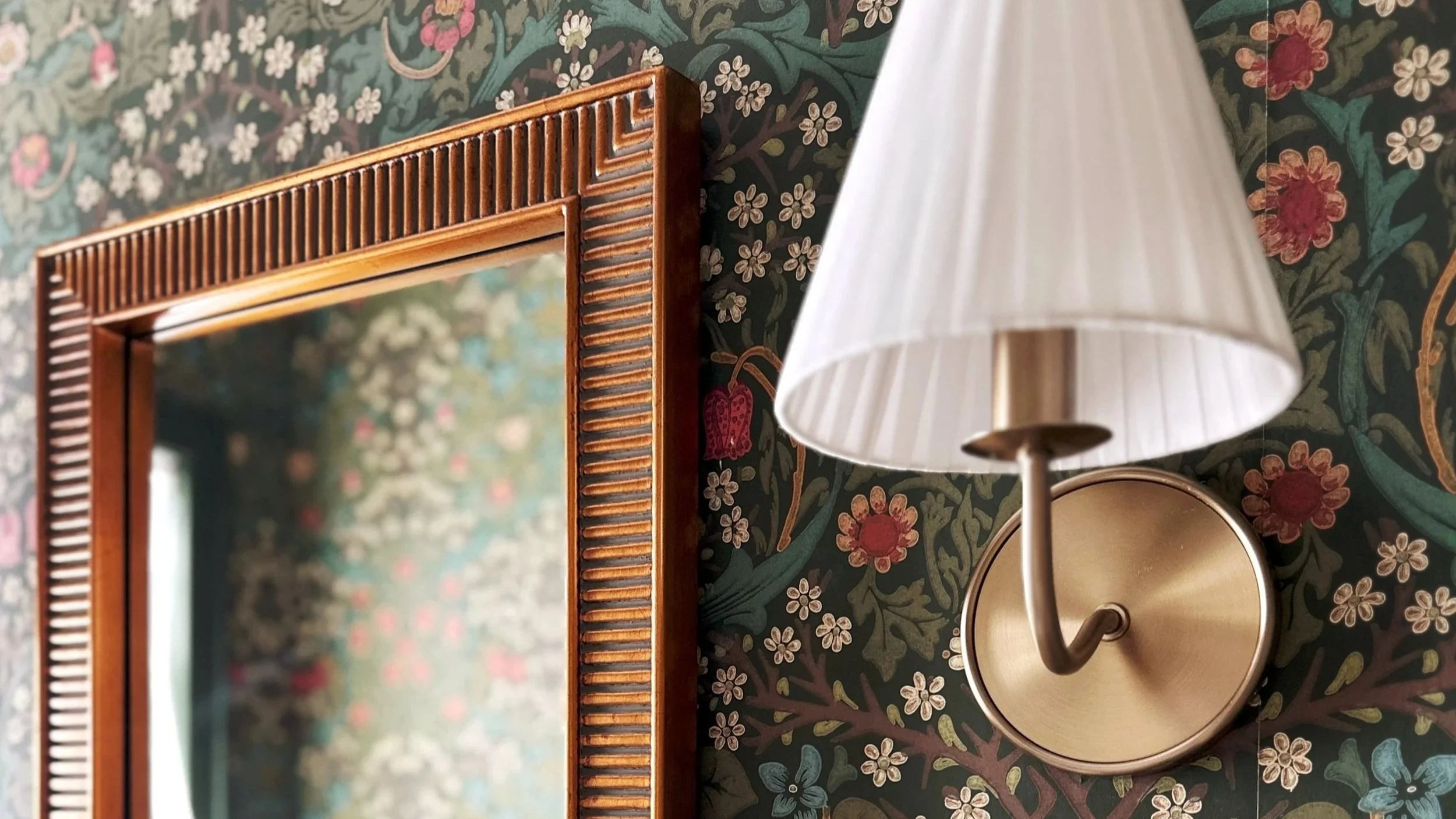 A decorative wall mirror with a carved wooden frame, a wall-mounted brass sconce with a white pleated lampshade, and floral wallpaper with a dark background, colorful flowers, and green leaves.