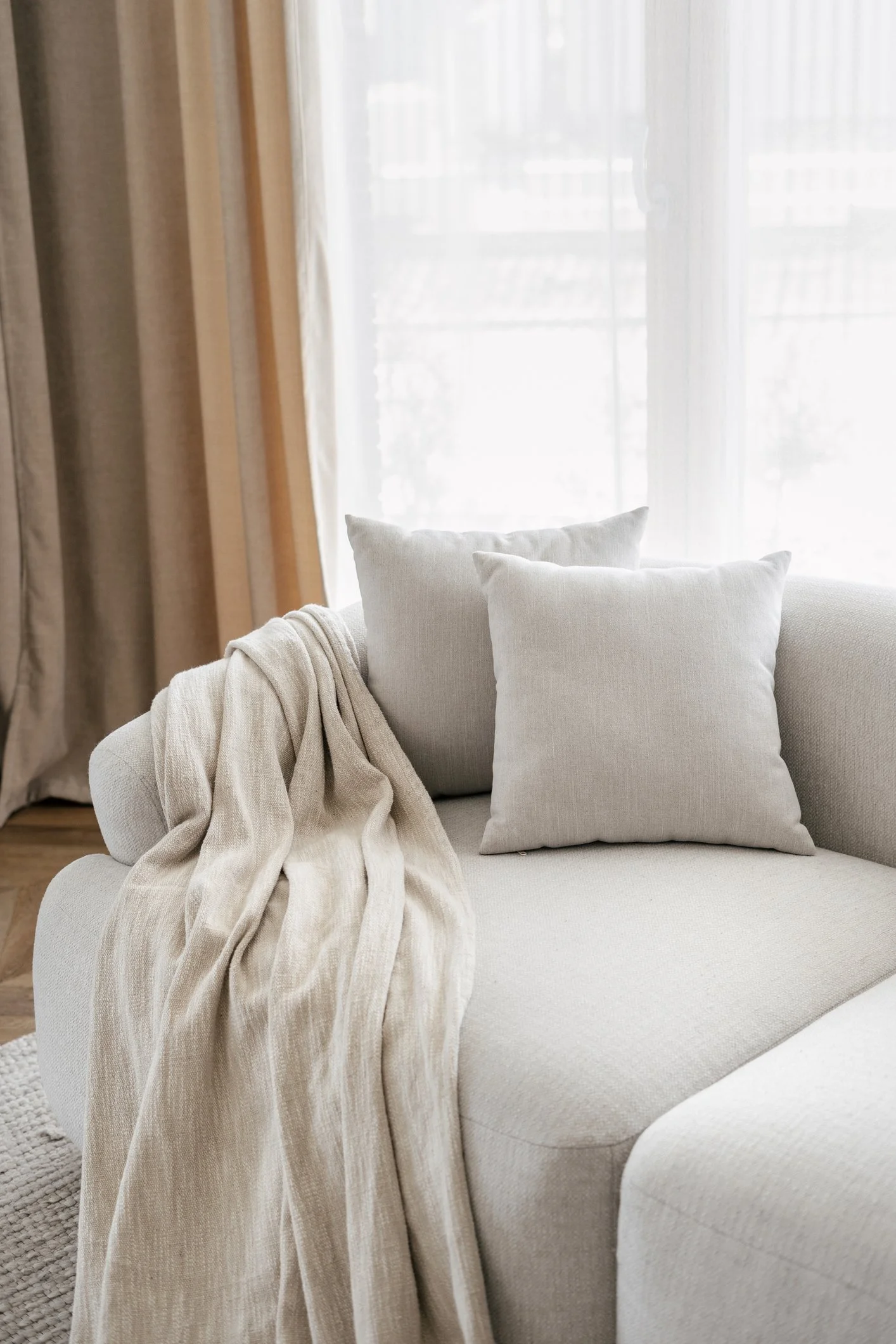 A beige sofa with two matching pillows and a cream-colored throw blanket in a living room with sheer curtains and a window