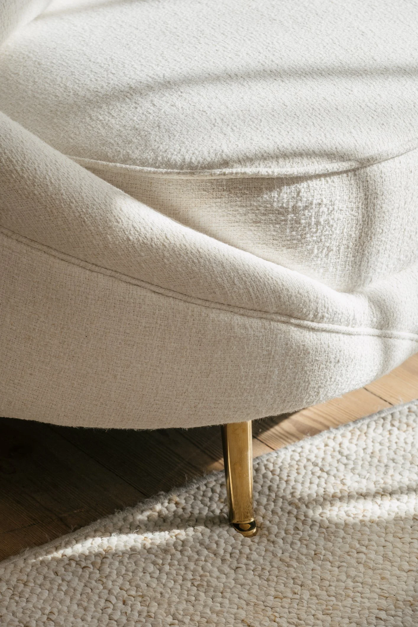 Close-up of a cream-colored upholstered chair with a gold leg sitting on a textured cream rug.