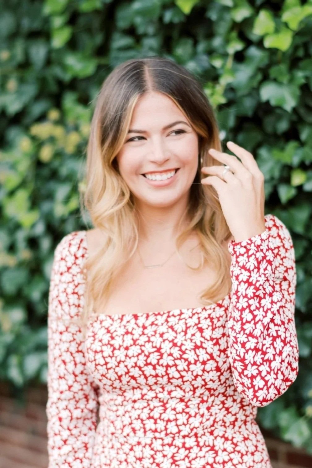 A young woman with wavy blonde hair, wearing a red dress with white floral pattern, smiling and touching her face with her right hand, outdoors with green foliage in the background.