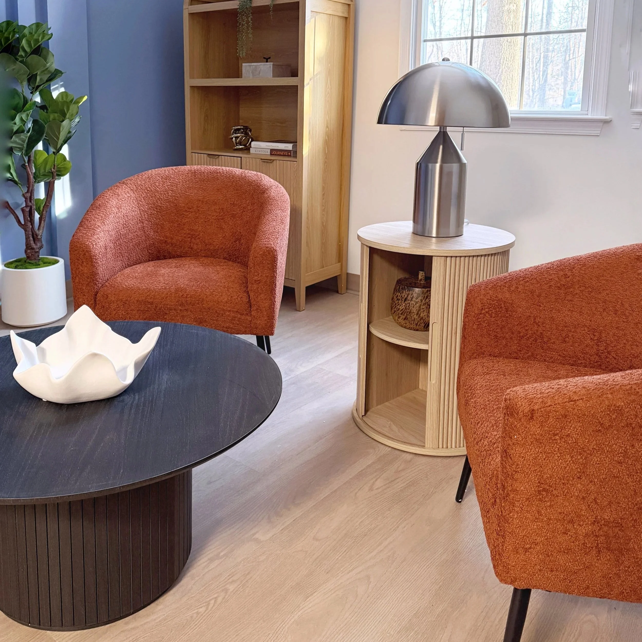 Living room with two orange armchairs, a black oval coffee table with a white decorative bowl, a wooden side table with a metallic lamp, a tall wooden shelf, and a potted plant.