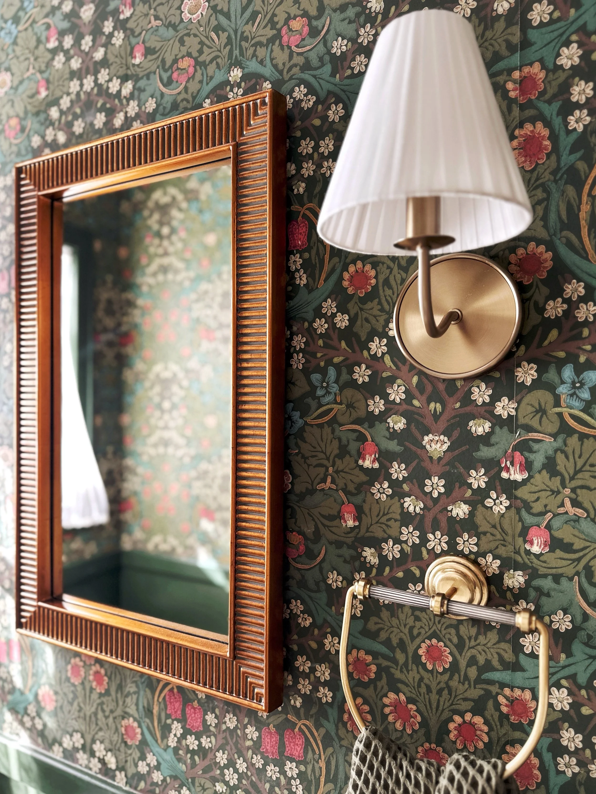 A decorative wall with a floral wallpaper, a wooden framed mirror, a wall-mounted lamp with a white shade, a brass towel holder with a patterned towel, all reflecting a window with a white curtain.