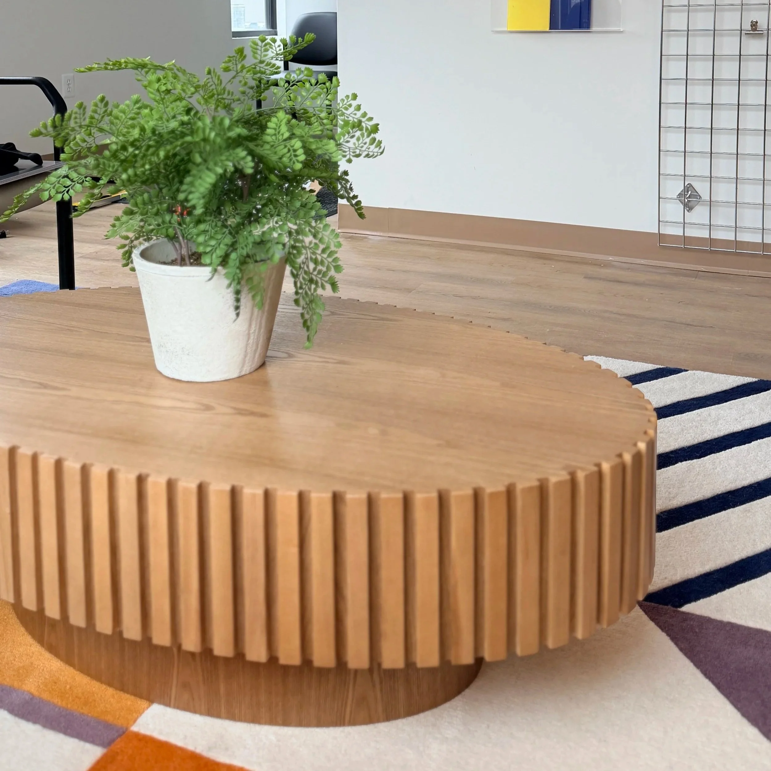 A round wooden coffee table with vertical slats around the edge is placed on a multicolored rug, and a potted green fern is on top of it. In the background, there is a white wall, hardwood floor, and part of a wire grid wall with some colored artwork.