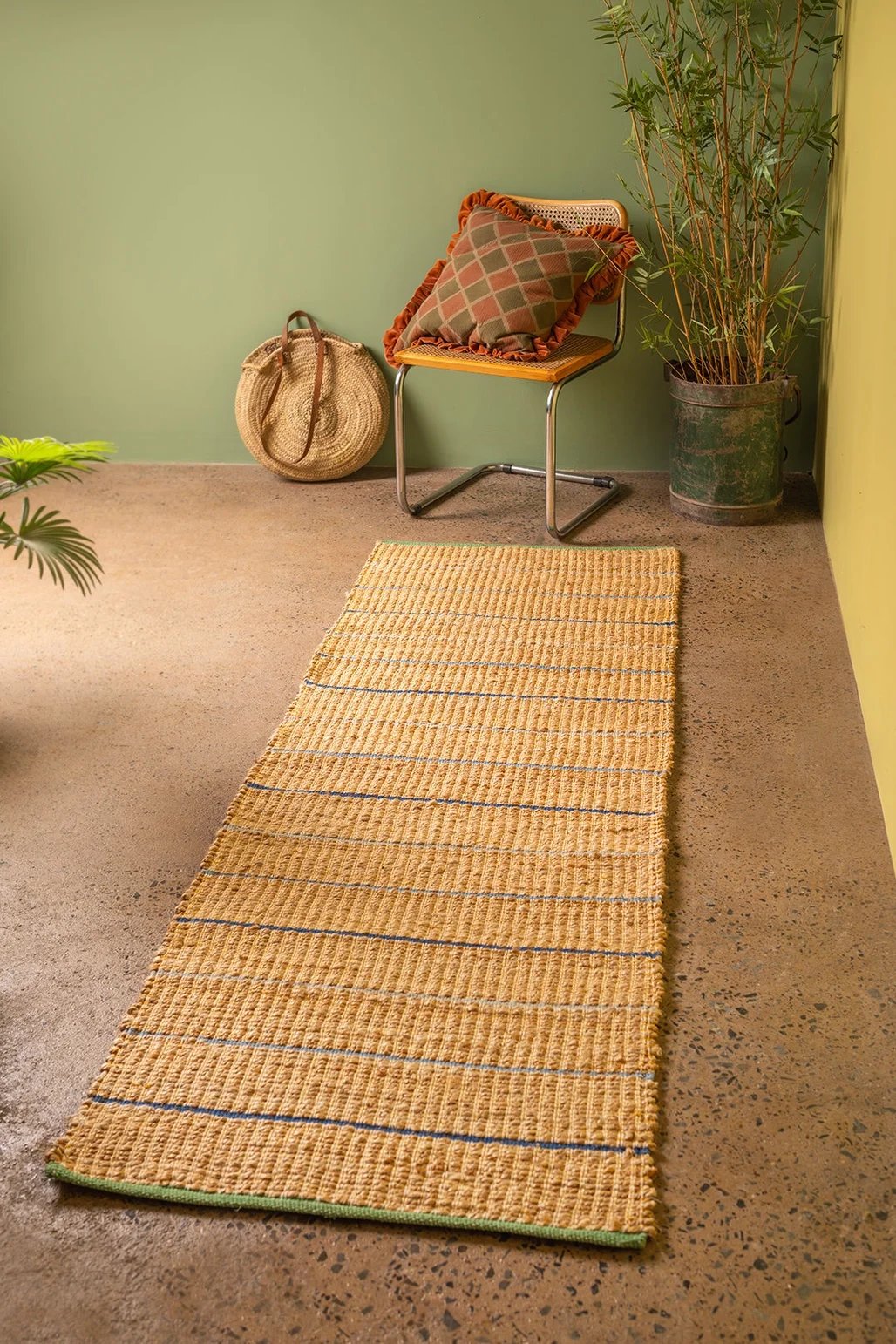 A room corner with a green wall, a beige textured rug on a brown floor, a chair with a plaid pillow, a large straw bag, and a tall potted plant.
