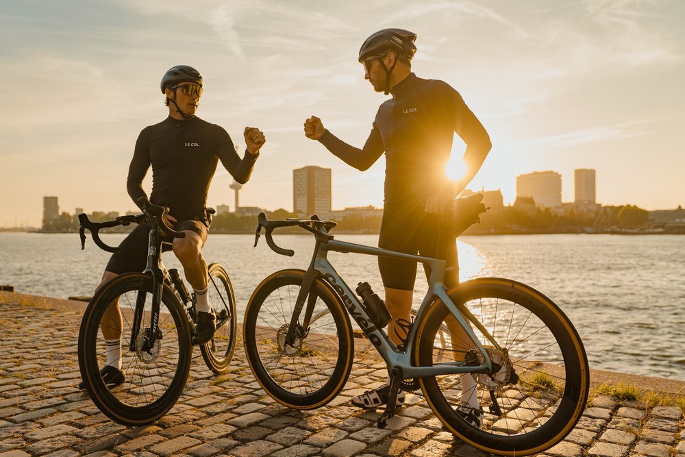 Twee wielrenners geven elkaar een boks aan de Maas in Rotterdam bij zonsondergang
