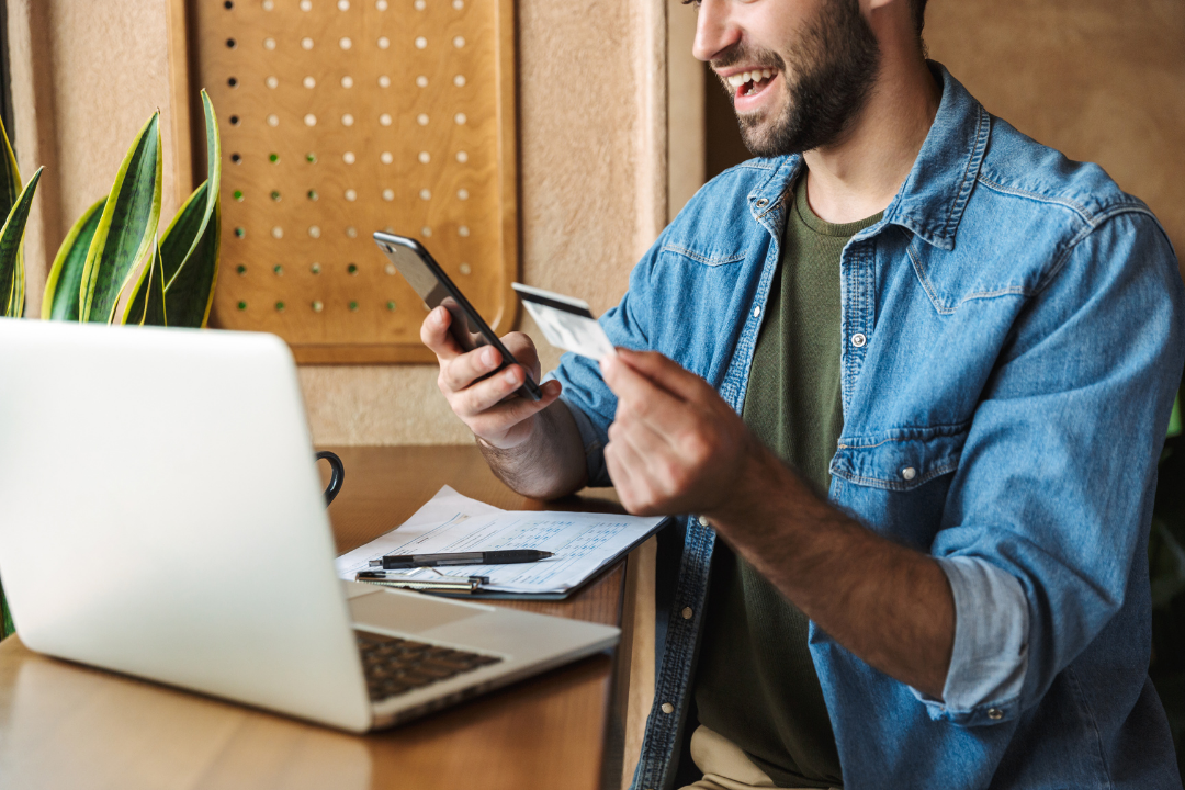 Man smiling and looking at his phone while sitting at a desk with a laptop, notepad, and pen, in a coffee shop or workspace.