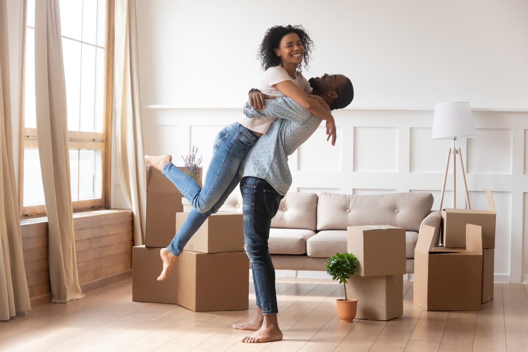 A happy couple moving into a new home with cardboard boxes. The man is lifting the woman in a piggyback ride as they smile and pose for the photo inside a well-lit living room.