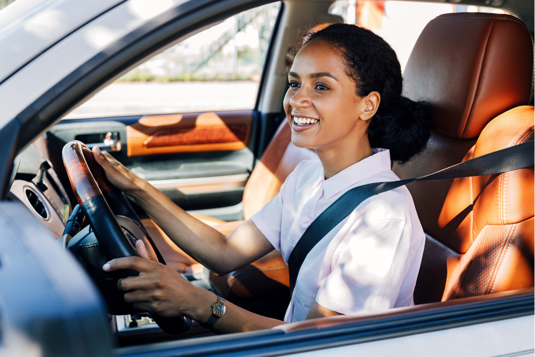 A woman with curly black hair and a white TB dress shirt sitting in the driver's seat of a car, smiling while holding the steering wheel.