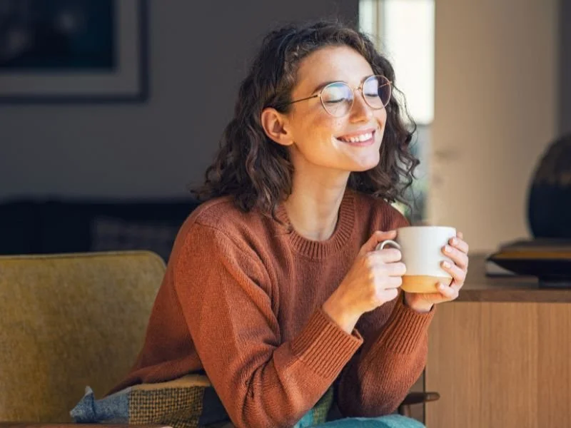A woman with curly hair and glasses smiles while holding a white mug, sitting in a cozy room.