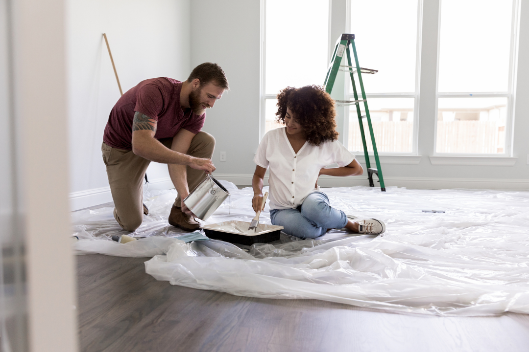 A man and a woman painting a room together. The man is kneeling, pouring paint, and the woman is sitting on the floor holding a paintbrush. The room has large windows and a step ladder.