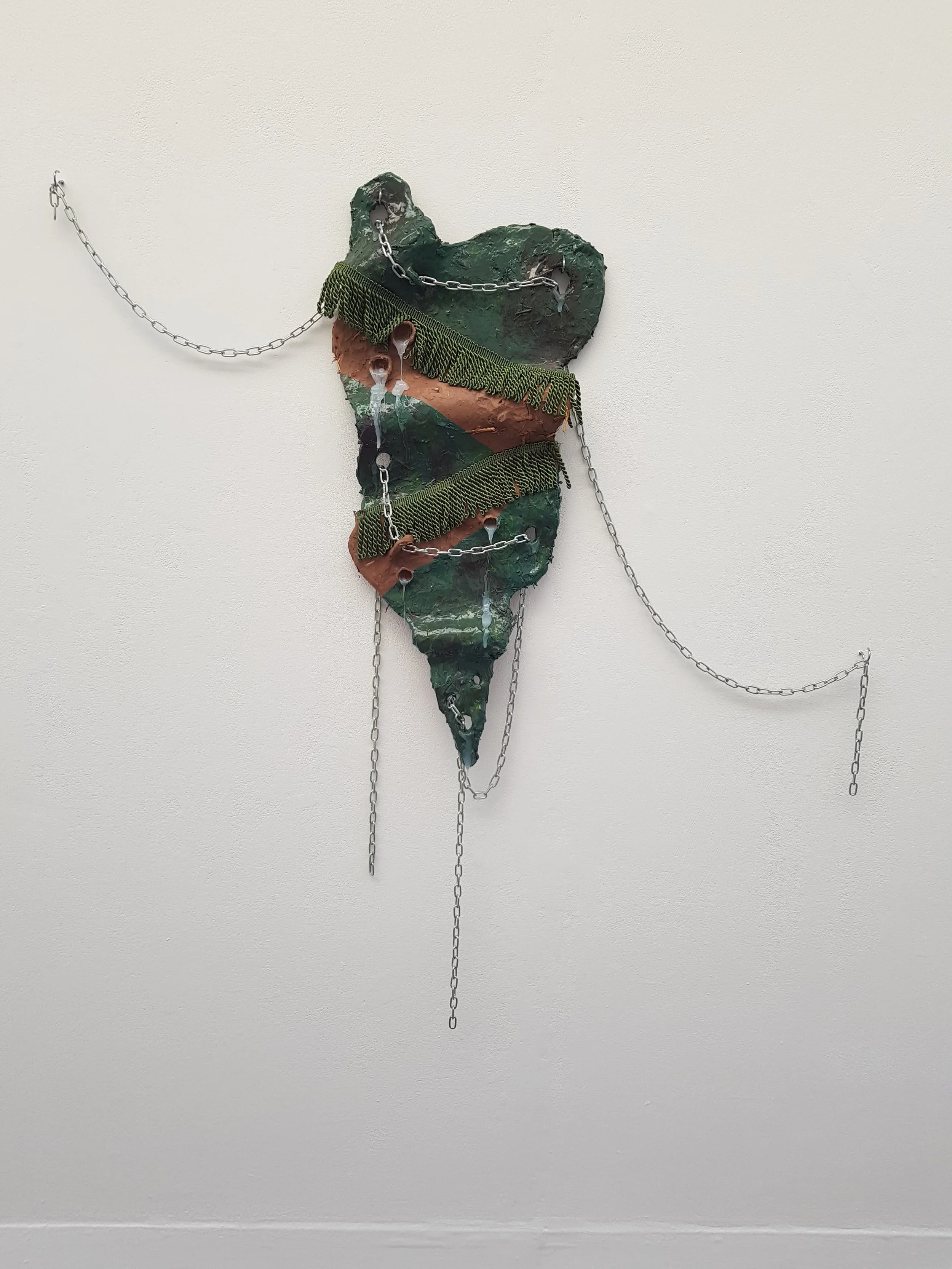 Mixed media art piece depicting a large, irregularly shaped object resembling a tooth, adorned with green and brown paint, fabric, and chains, mounted on a white wall.
