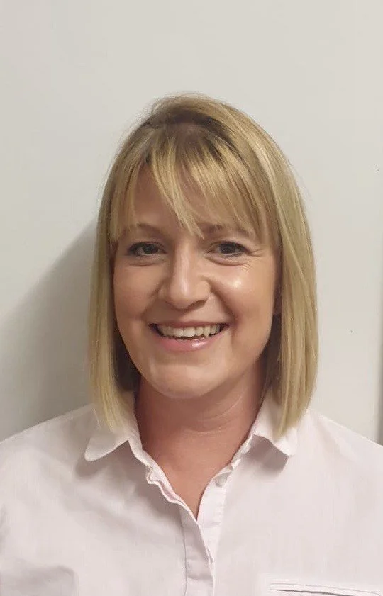A woman with blonde hair and a white collared shirt, smiling against a plain white wall.