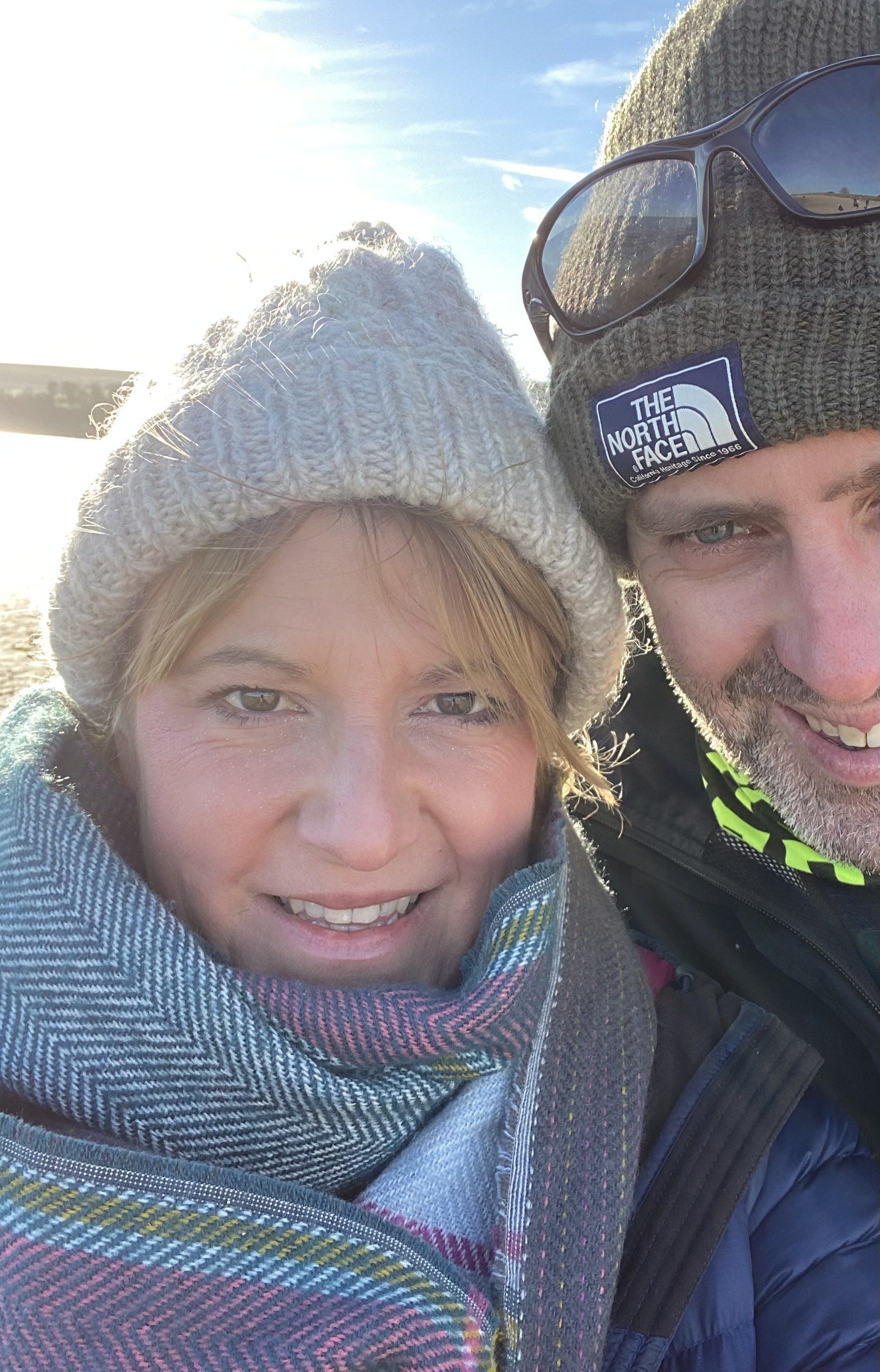 Close-up of a smiling woman and man outdoors in winter attire, wearing beanies and jackets, with the sky and sunlight behind them.