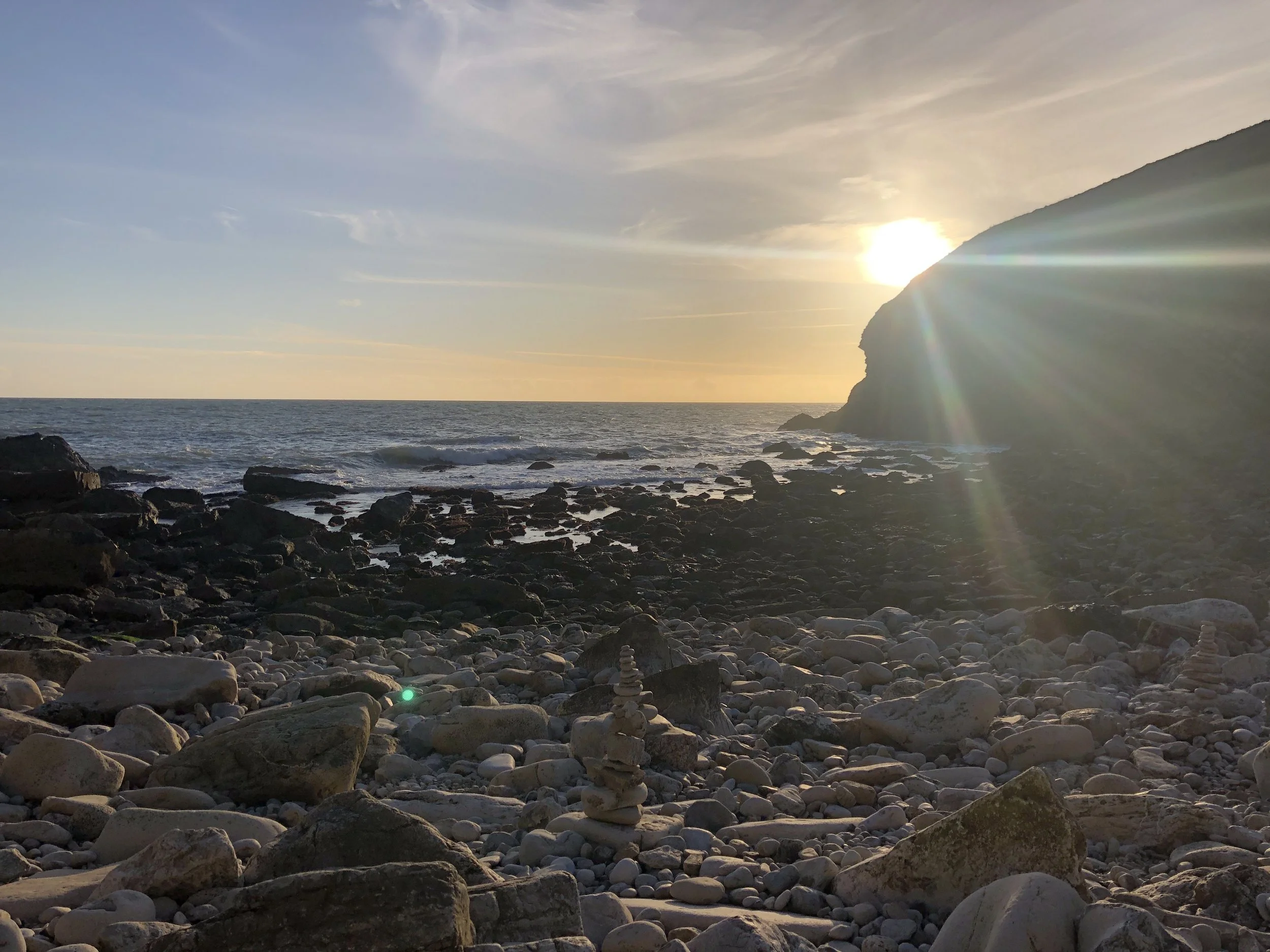 A rocky beach at sunset with a large cliff on the right, the sun setting behind it, casting rays of light over the scene, and stacks of rocks balanced on the beach.