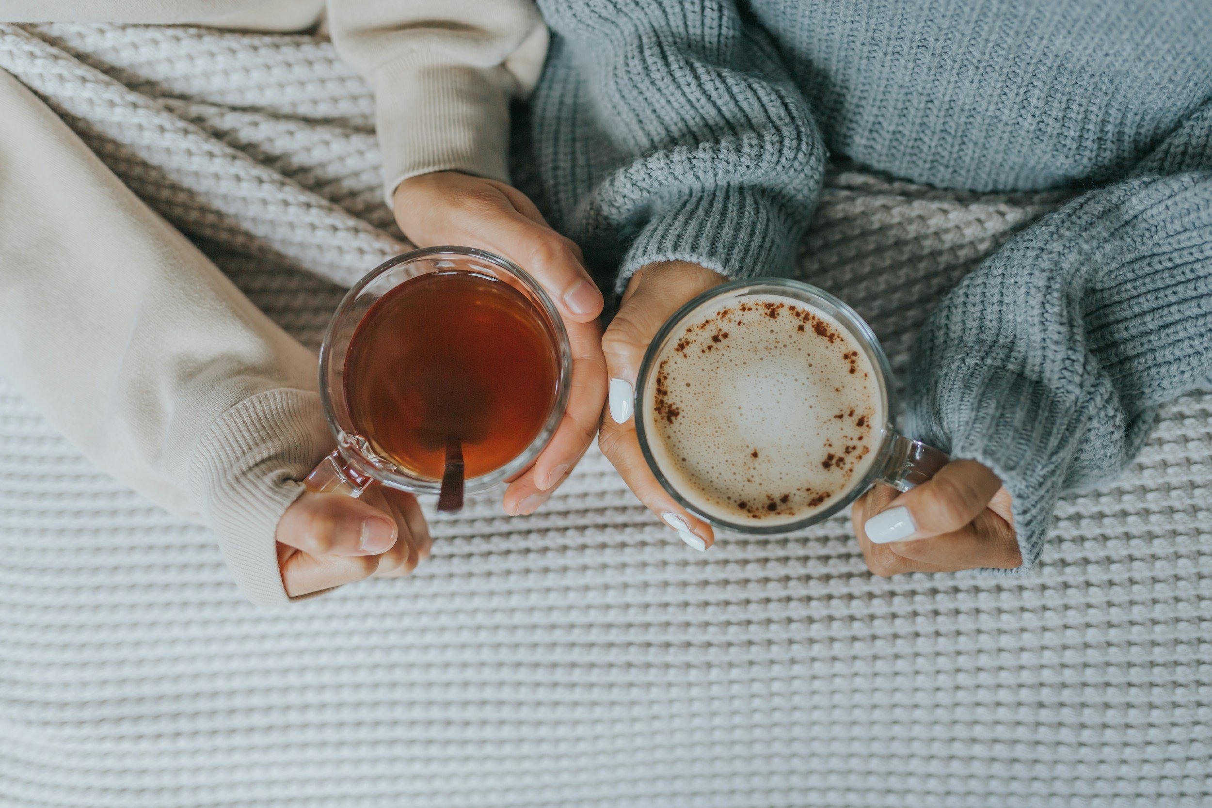 Two people holding cups with hot beverages, one with tea and the other with a frothy drink, sitting on a textured blanket.