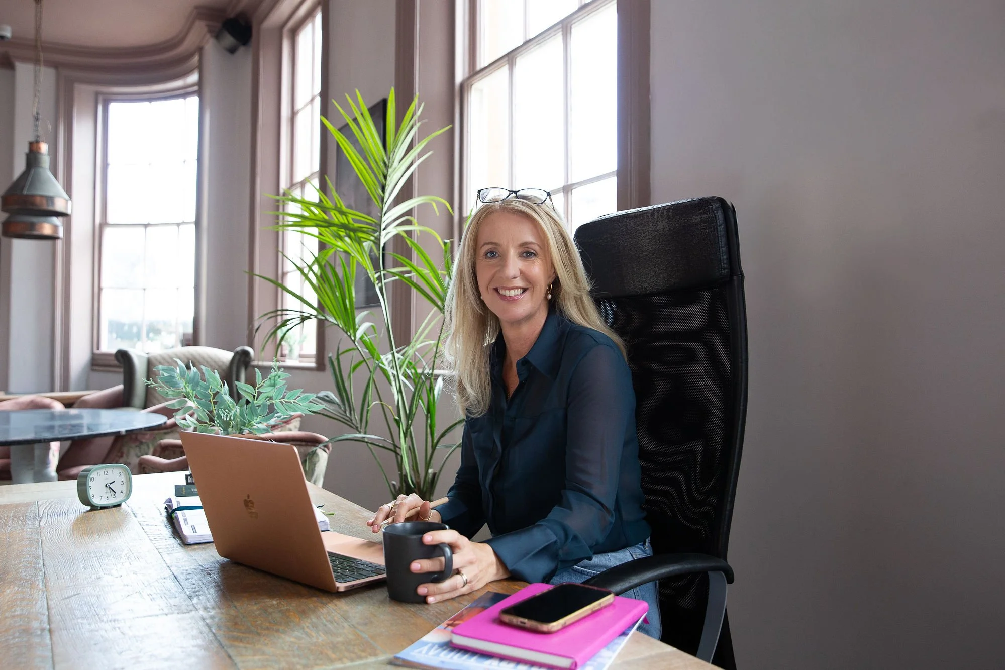 Claire dale , therapist  blonde hair sitting at a desk with a laptop, holding a coffee mug and smiling. The office is bright with large windows and green plants.
