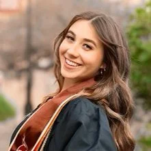 Smiling young woman with long brown hair outdoors, wearing a dark jacket and a brown shirt.