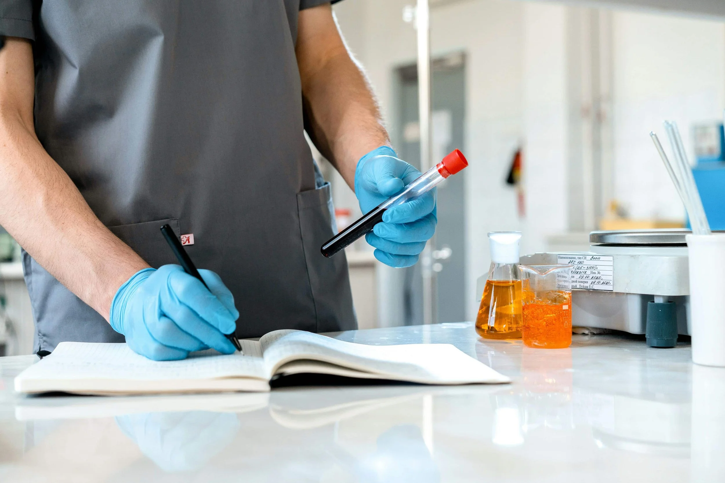 A lab technician in blue gloves holding a blood collection tube while recording notes in a laboratory notebook