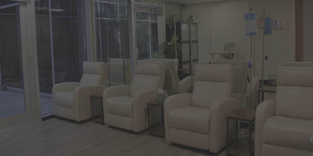 Interior of a modern healthcare facility with beige recliner chairs arranged in a row, small wooden side tables, medical equipment, and large glass doors leading to an outdoor area.