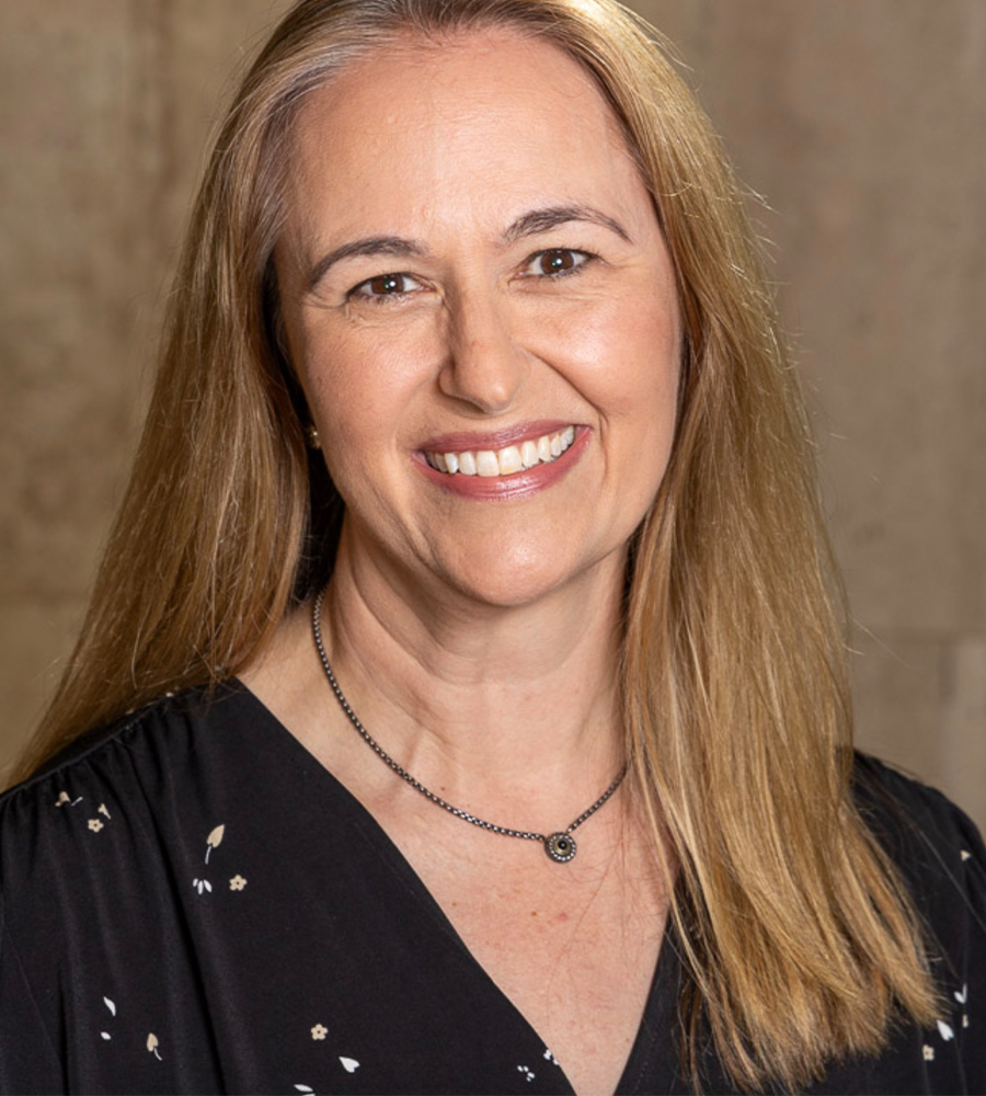 Smiling woman with long blonde hair, wearing a black dress with small white floral pattern and a black beaded necklace, posing in front of a neutral background.