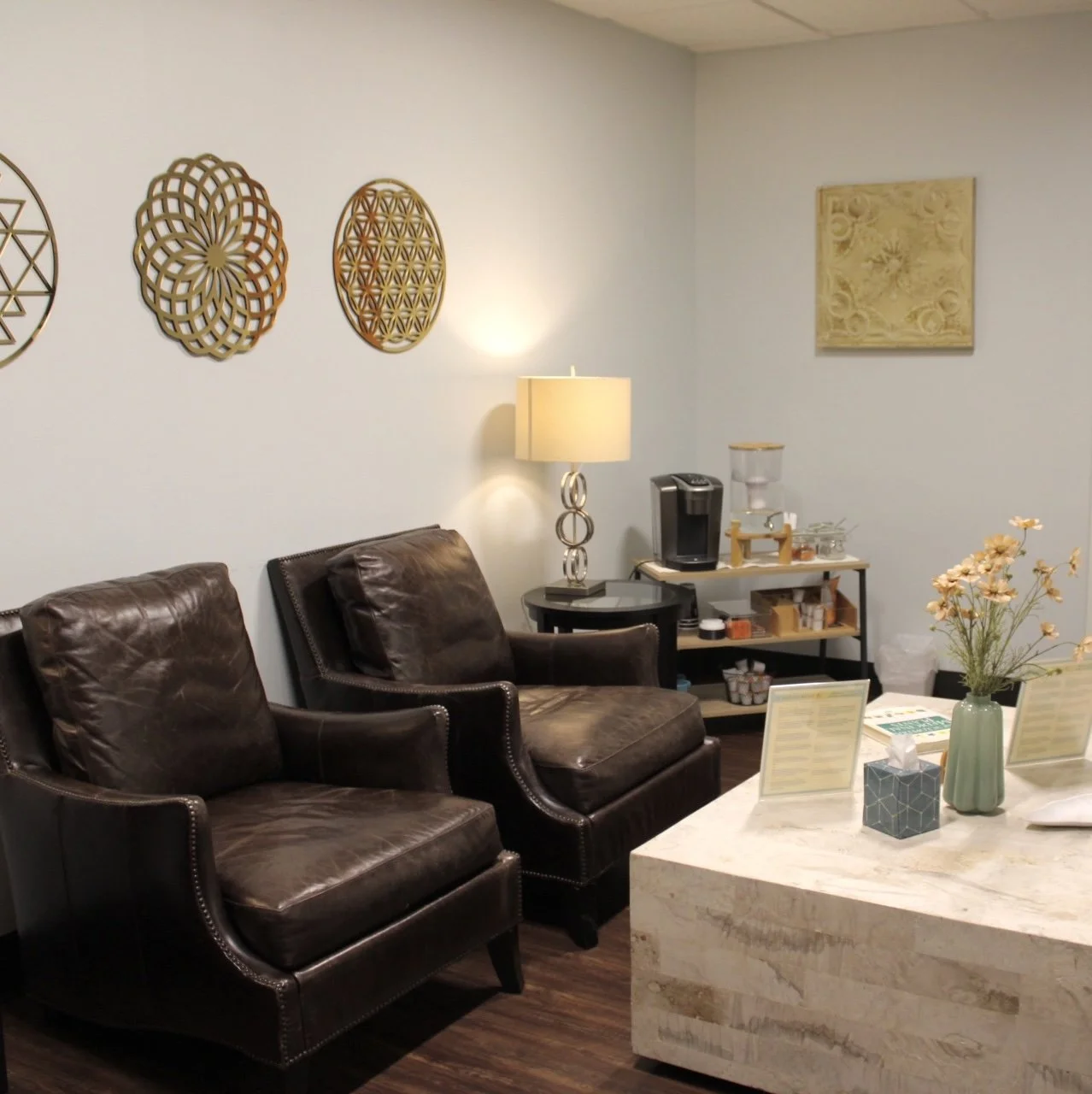 A waiting room with two brown leather armchairs, a lamp, a coffee maker, a water dispenser, a table with flowers, tissue box, and informational pamphlets, and decorative wall hangings.
