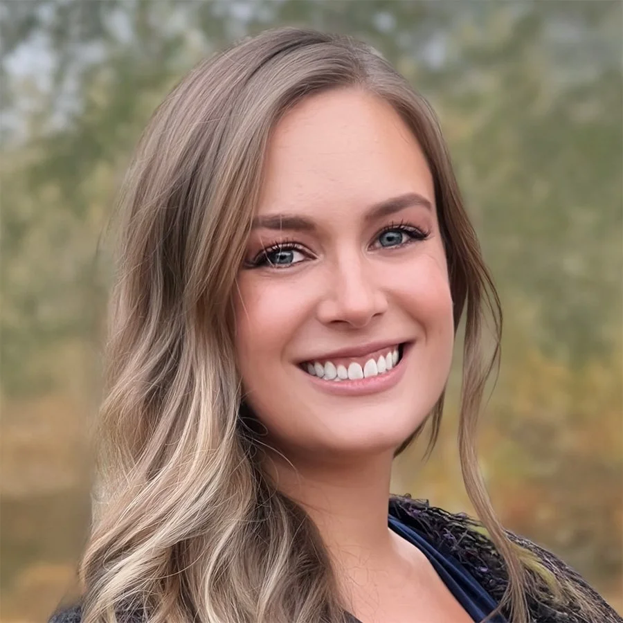 A young woman with long, wavy blonde hair smiling outdoors with blurred trees in the background.