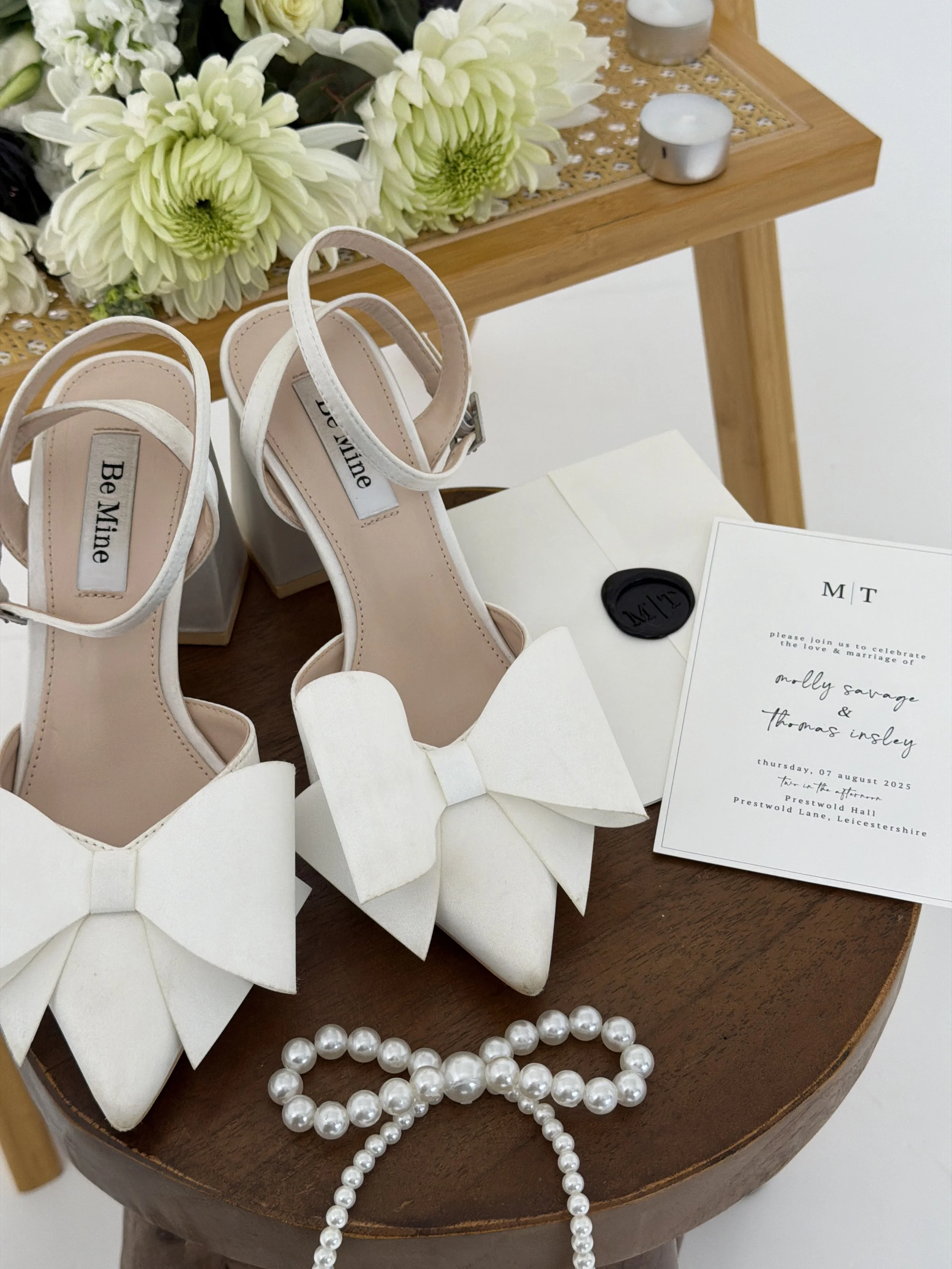White high-heeled shoes with large bows, a pearl necklace, a wedding invitation, and floral decorations on a wooden table.