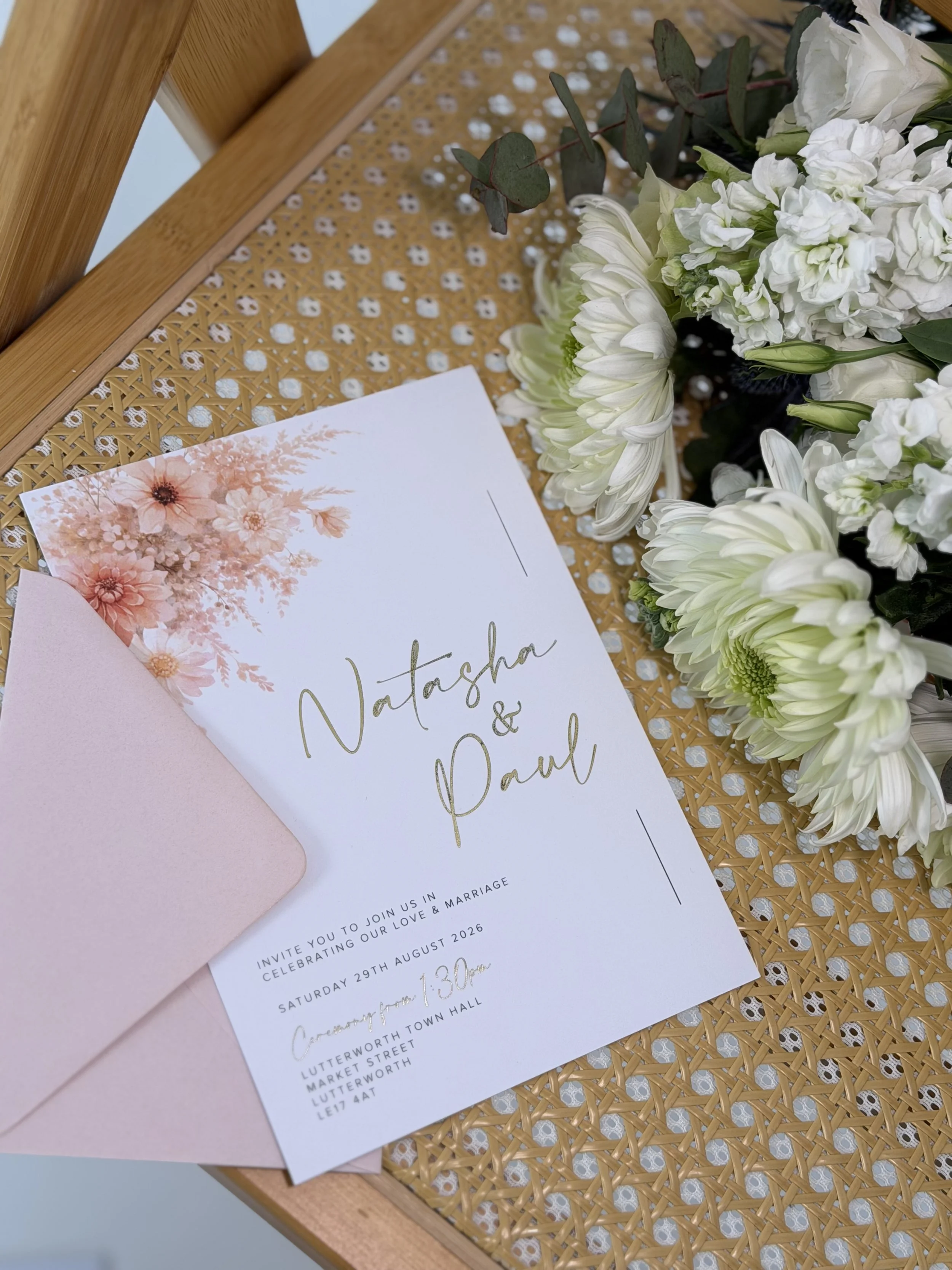 Wedding invitation card for Natasha and Paul with floral design, placed on a woven surface next to a bouquet of white flowers.