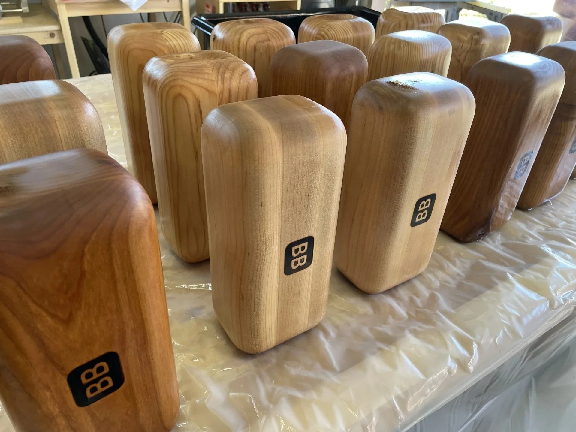 Multiple wooden blocks with rounded edges, some with a black 'BB' logo, arranged on a plastic-covered table, in what appears to be a woodworking or crafting environment.