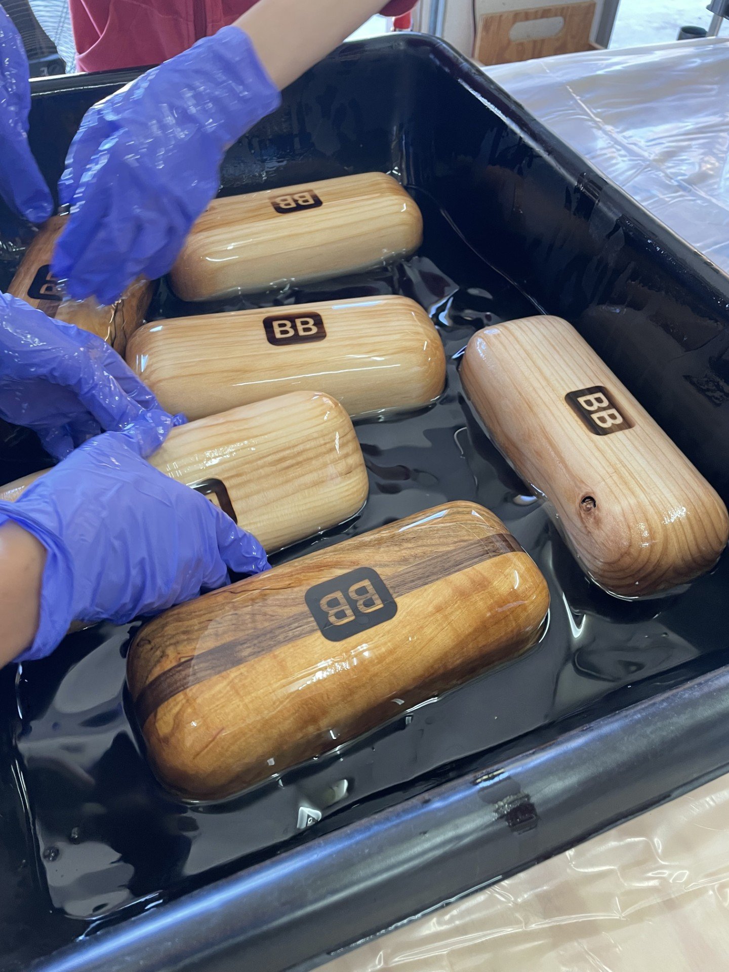 Multiple wooden objects with the letters 'BB' stamped on them floating in a black container filled with water, with a pair of hands wearing blue gloves handling one of the objects.