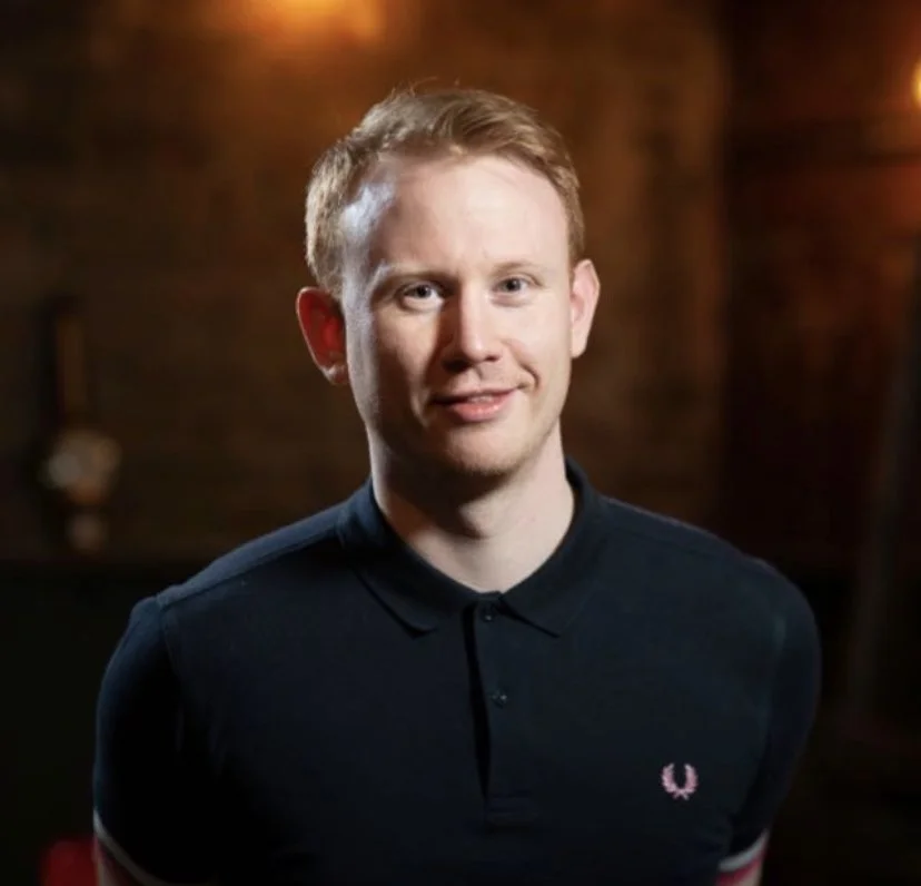 A young man with short red hair and fair skin smiling, wearing a black polo shirt with a small pink logo, standing in a dimly lit indoor setting.