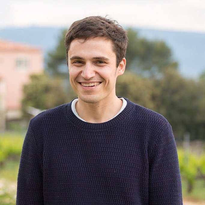 A young man with short brown hair smiling outdoors in front of trees and a building, wearing a navy blue sweater.