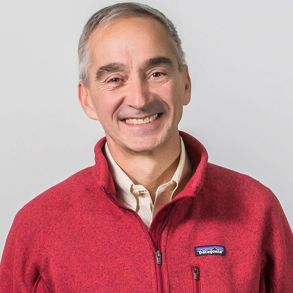 A smiling middle-aged man with gray hair wearing a red Patagonia jacket over a beige collared shirt against a plain gray background.