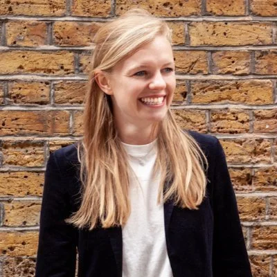 A smiling woman with long blonde hair, wearing a black jacket over a white shirt, standing in front of a brick wall.