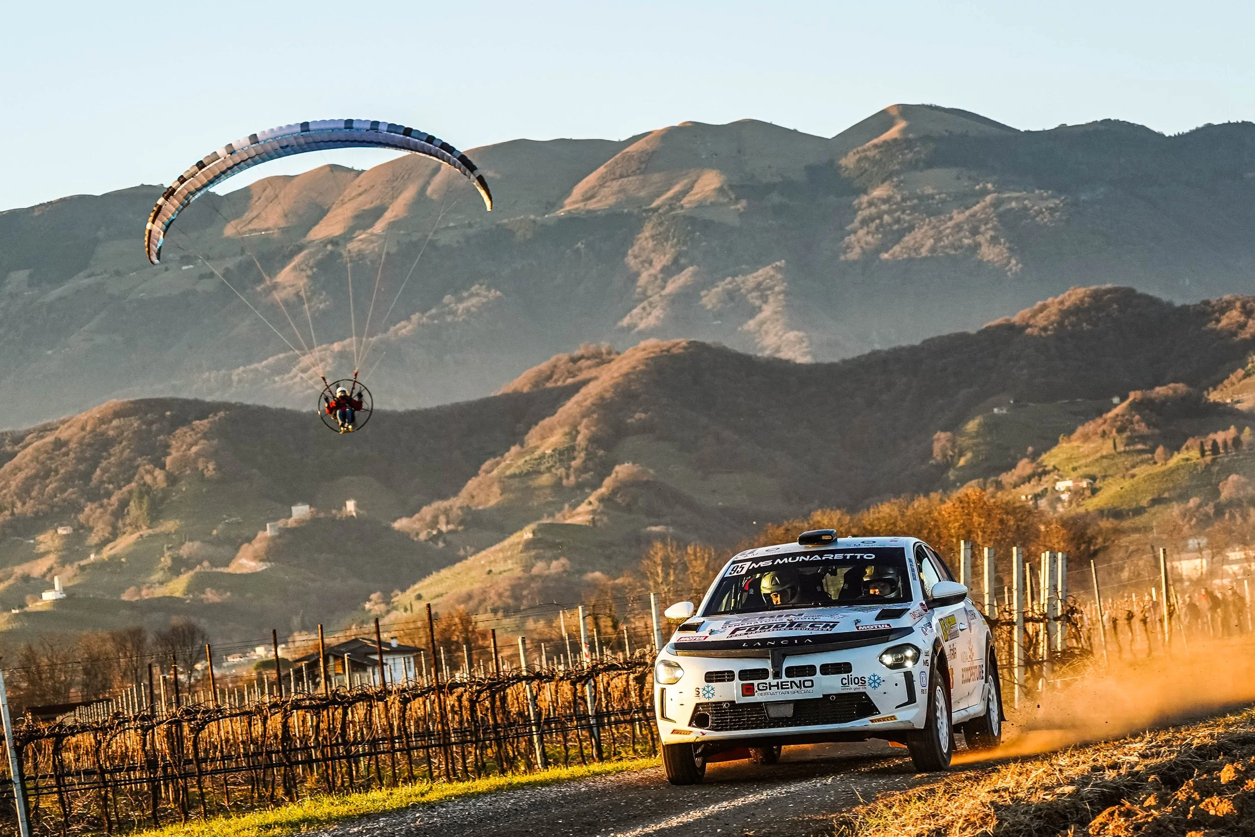 Auto da rally che corre lungo una strada di campagna con vigneti, sullo sfondo montagne e un pilota di parapendio in volo.