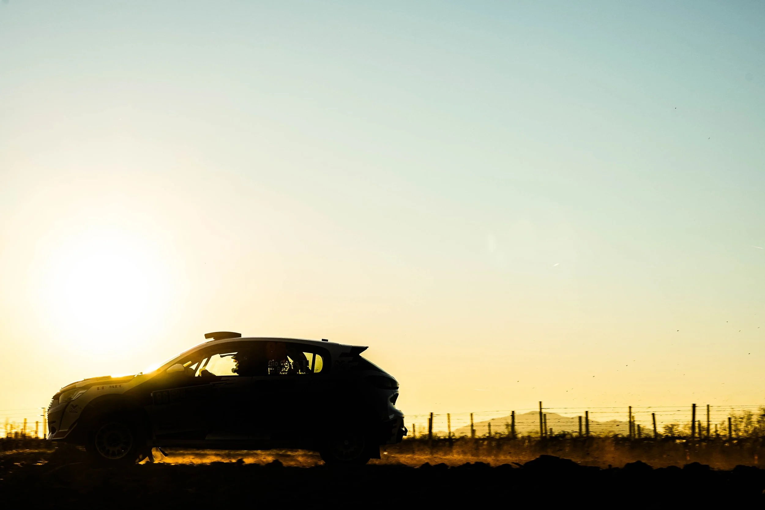Silhouette di un'auto da corsa che percorre una strada in campagna al tramonto, con il cielo chiaro e una recinzione in lontananza.