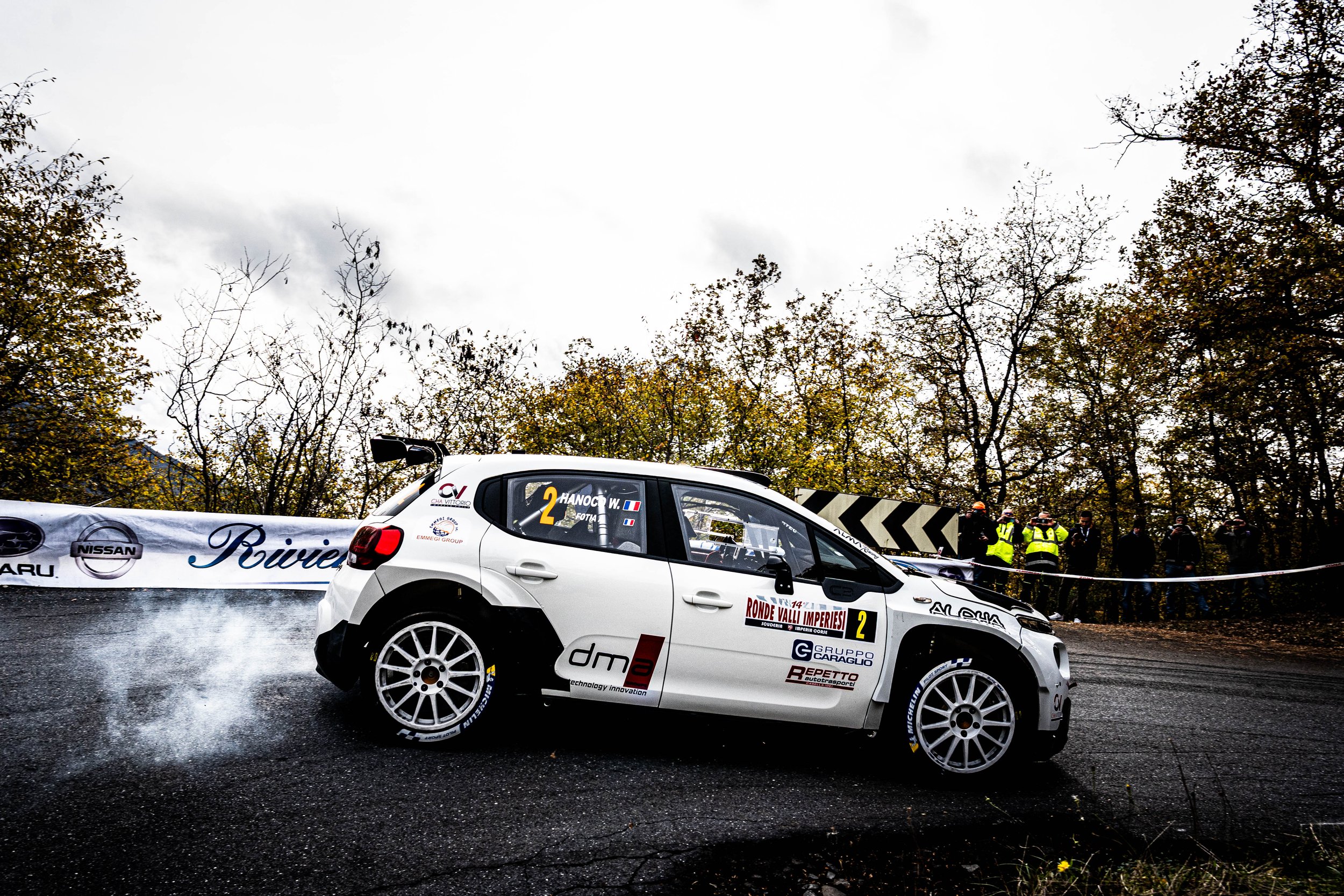 Auto da rally che scivola su una strada asfaltata durante una competizione, con spettatori e alberi sullo sfondo.
