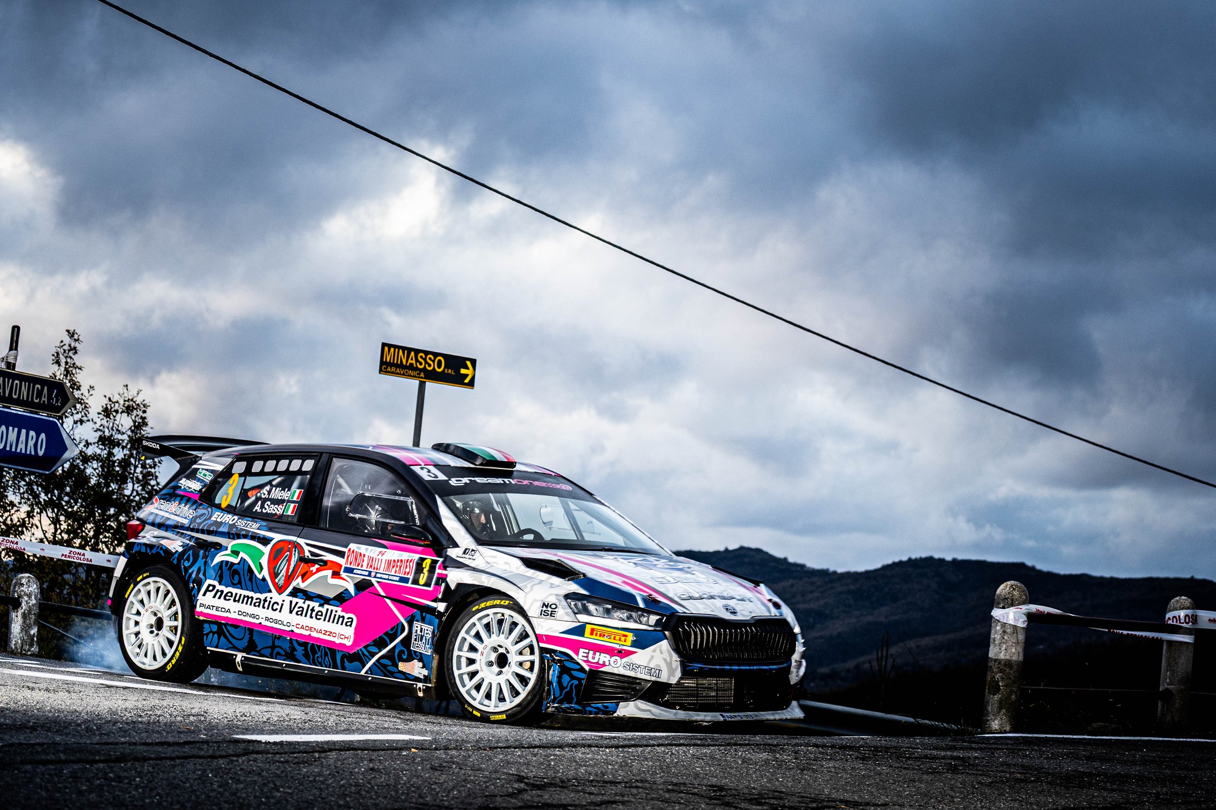 Auto da rally con colori vivaci e sponsor, in una giornata nuvolosa, lungo una strada di montagna con cartelli stradali e paesaggi collinari sullo sfondo.