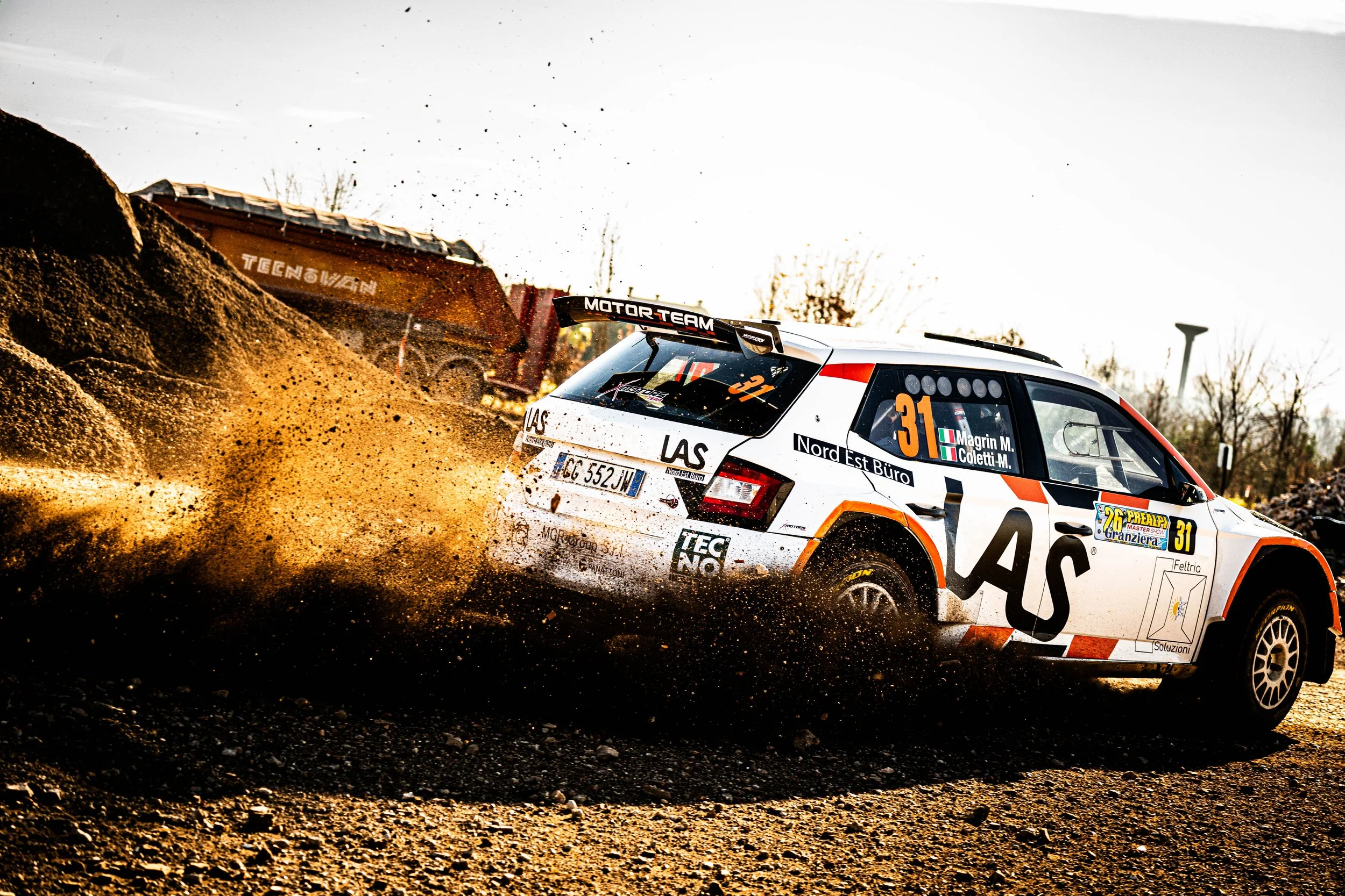 Auto da rally bianca con grafiche nere arancioni e il numero 31 che attraversa un terreno di ghiaia sollevando polvere e pietre durante una gara