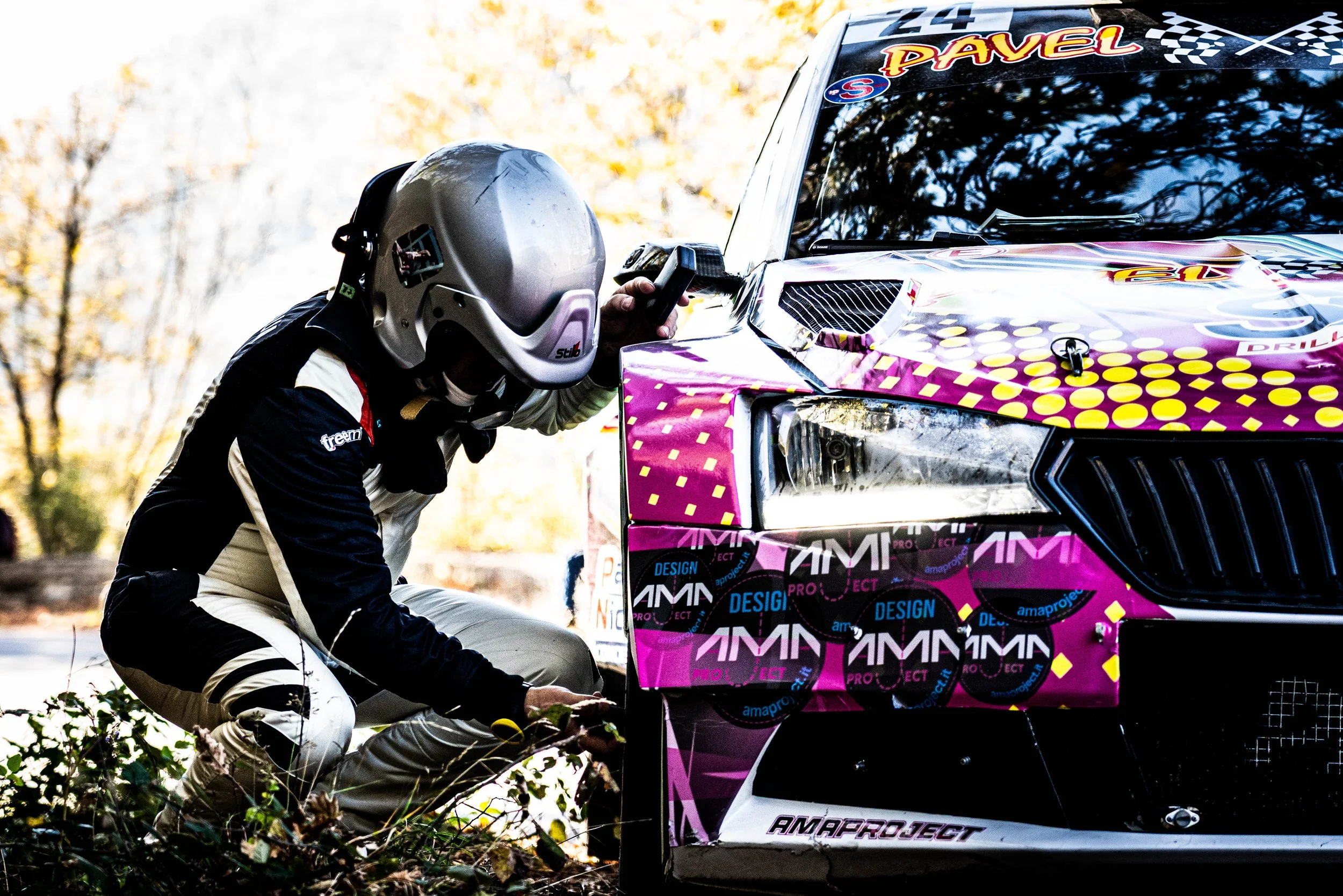 Un pilota di rally con casco appoggiato vicino a una macchina da rally decorata con colori rosa, viola e gialli, che si trova in un'area naturale con alberi autunnali sullo sfondo.