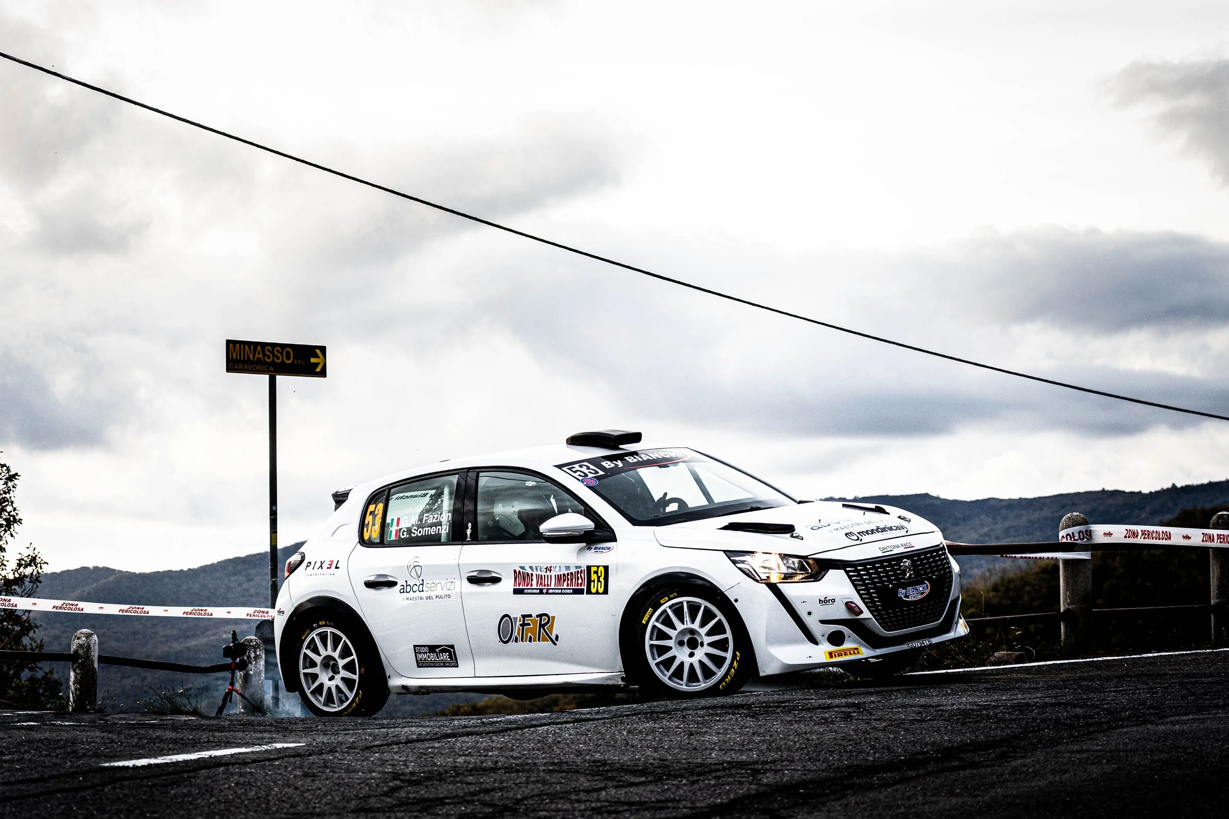 Auto da rally bianca sulla strada con sfondo montano e cielo nuvoloso.