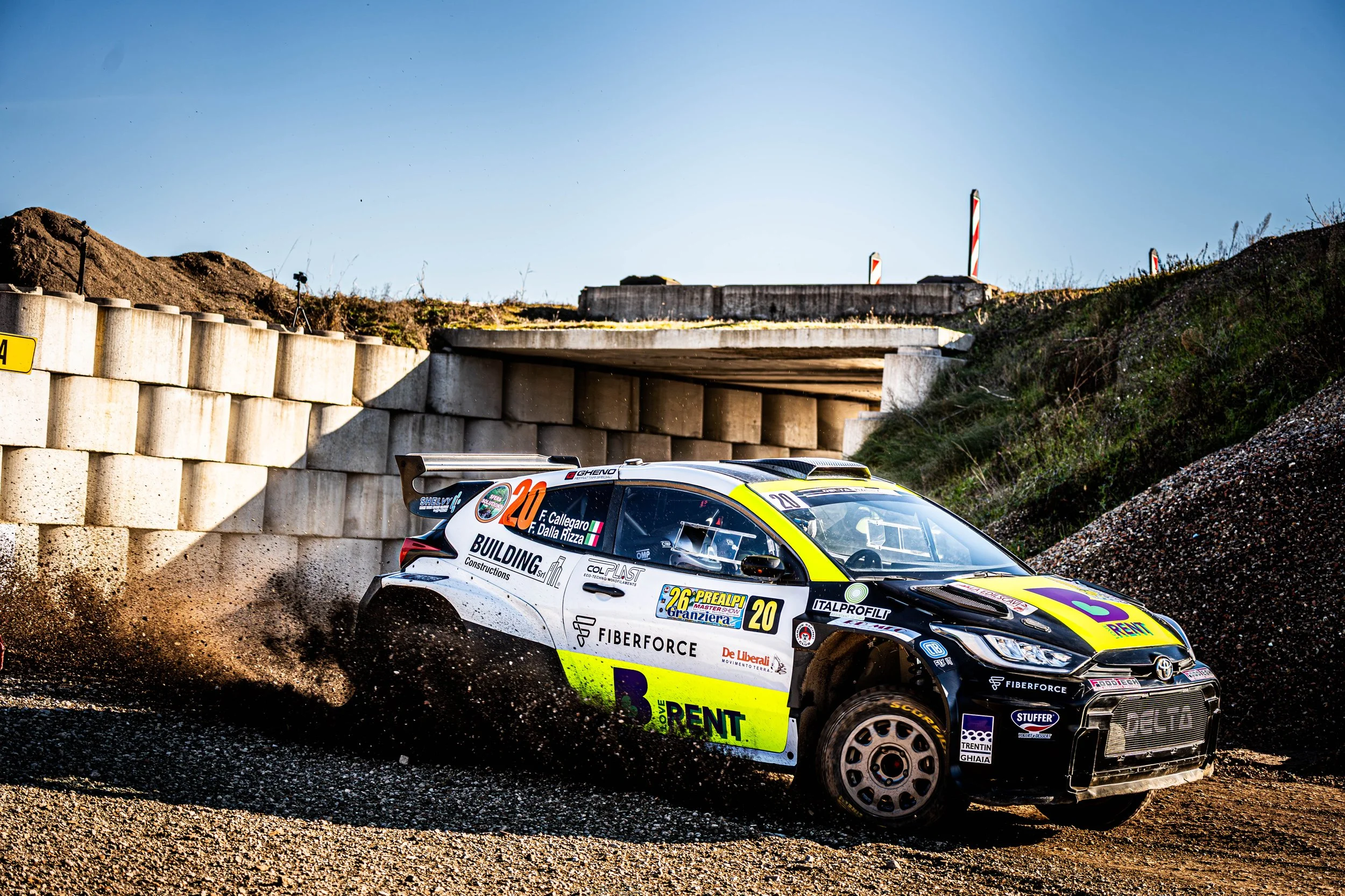 Un'auto da rally che sfreccia su un percorso sterrato, sollevando polvere, sotto un ponte durante una corsa.