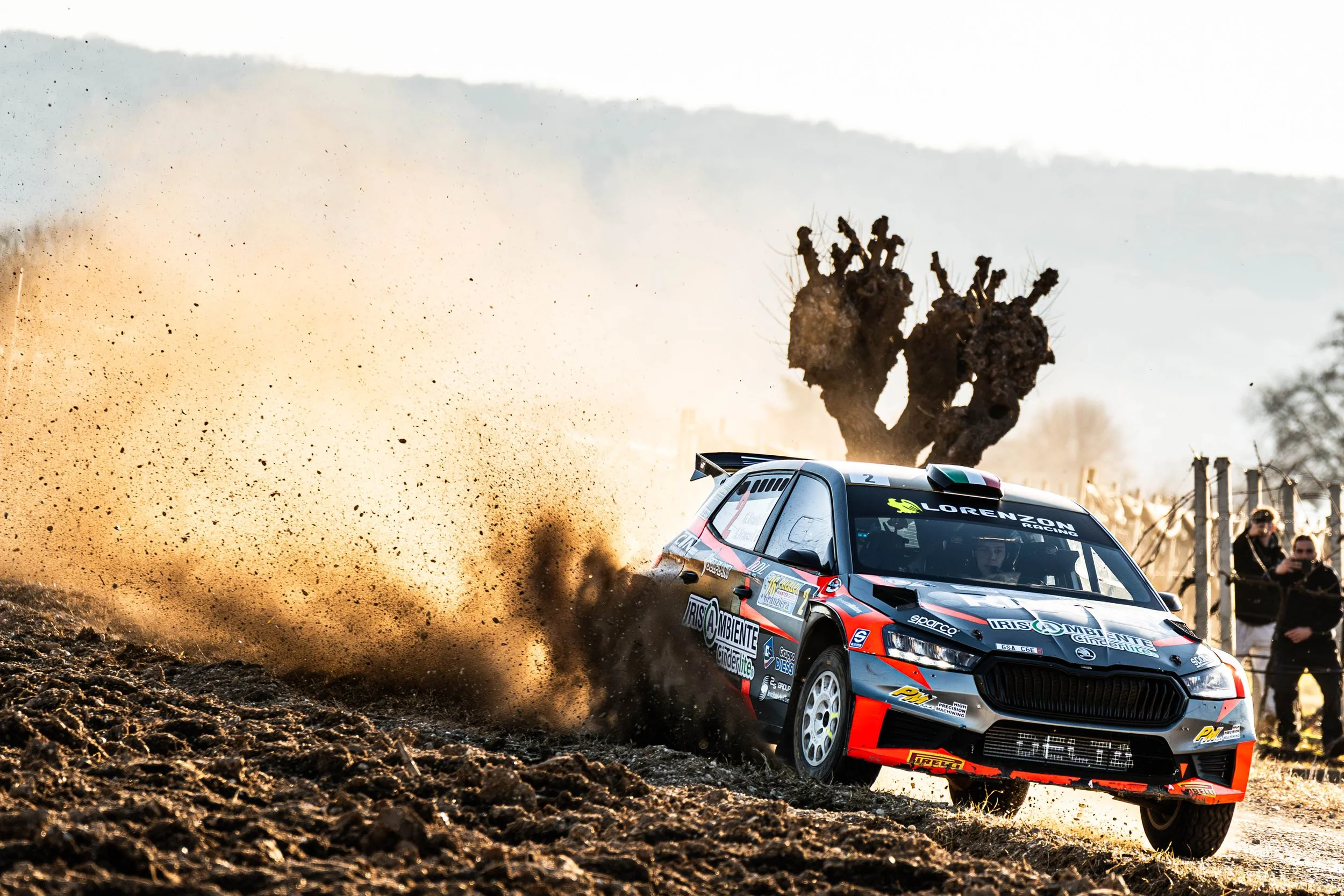 Auto da rally in curva su strada sterrata, solleva polvere e ciottoli, con due persone che fotografano sullo sfondo e un albero potenziato