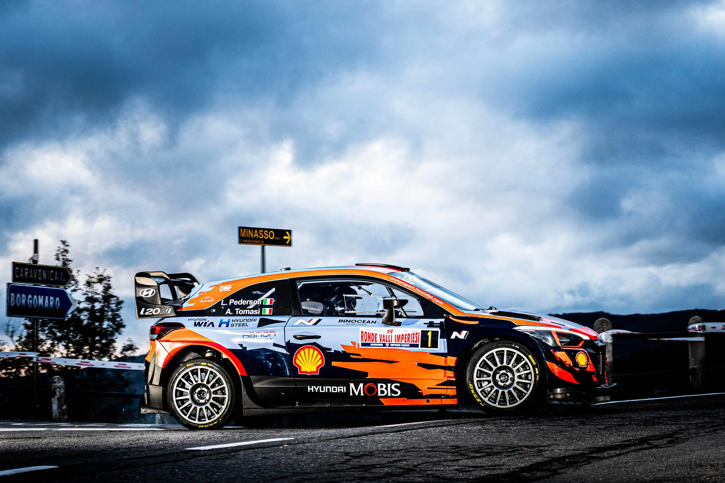 Auto da corsa rally Hyundai i20 N in livrea arancione, nero e bianco, con sponsor Shell e Hyundai Mobis, in curva su strada con cartelli di direzione e cielo nuvoloso sullo sfondo.