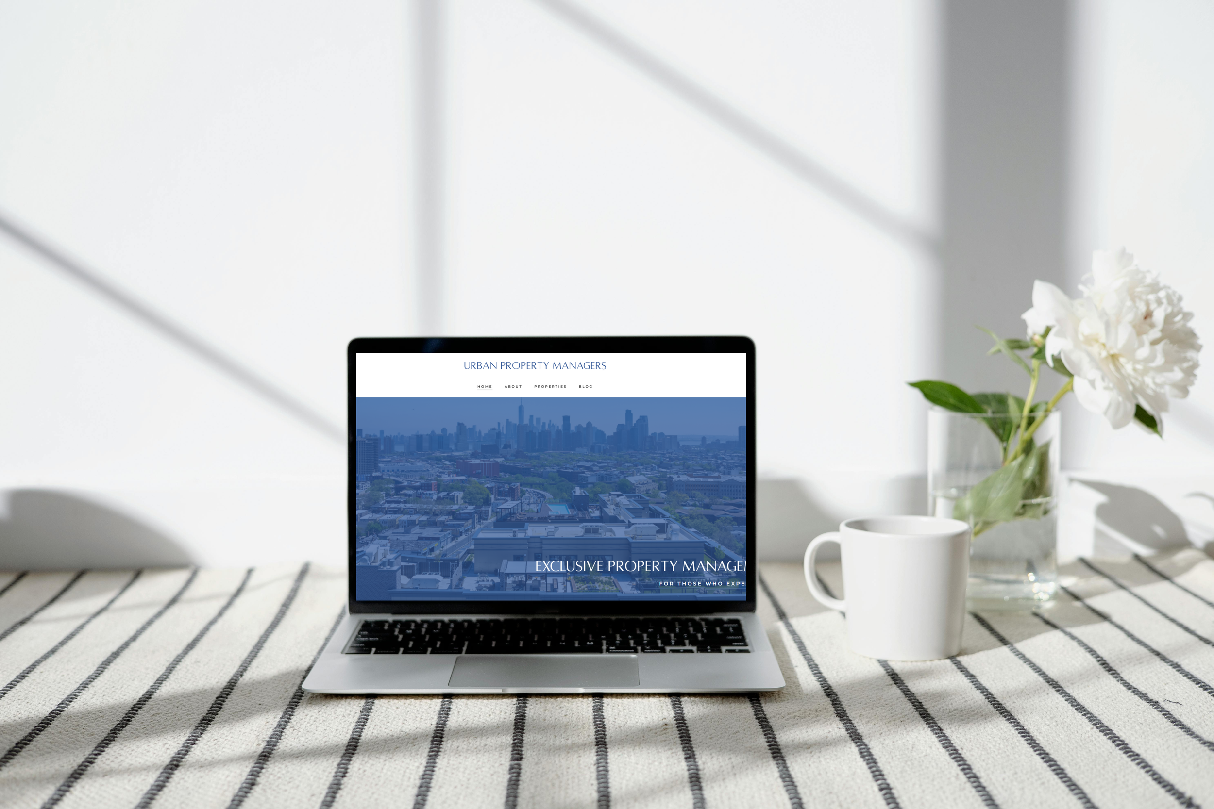 Laptop on a striped table showing a website for urban property managers, next to a white coffee mug and a glass vase with white flowers, in a bright room with sunlight.