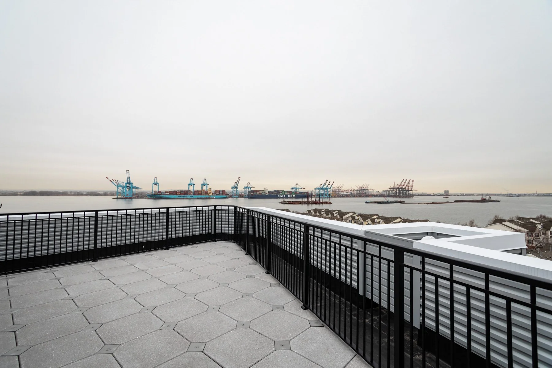 View from a rooftop terrace overlooking a harbor with cargo cranes and ships on an overcast day.