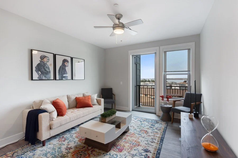 Living room with a white sofa, two orange pillows, a black armchair, a sliding door leading to a balcony, a wooden TV stand with a TV, and a colorful area rug.