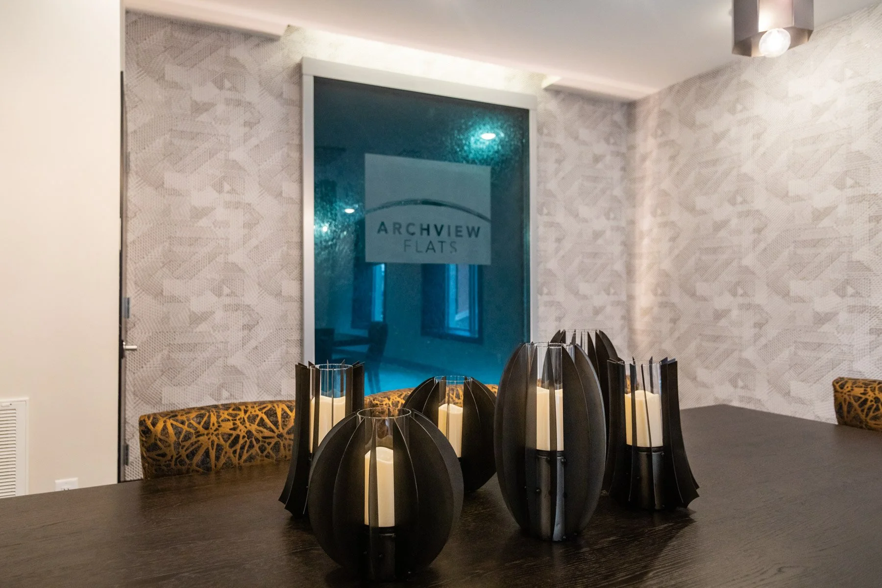 A dark wooden table with abstract black and white candle holders containing unlit white candles, set in a modern interior with a patterned white wall, patterned upholstered chairs, and a glass door with "Archview Flats" written on it in the background.