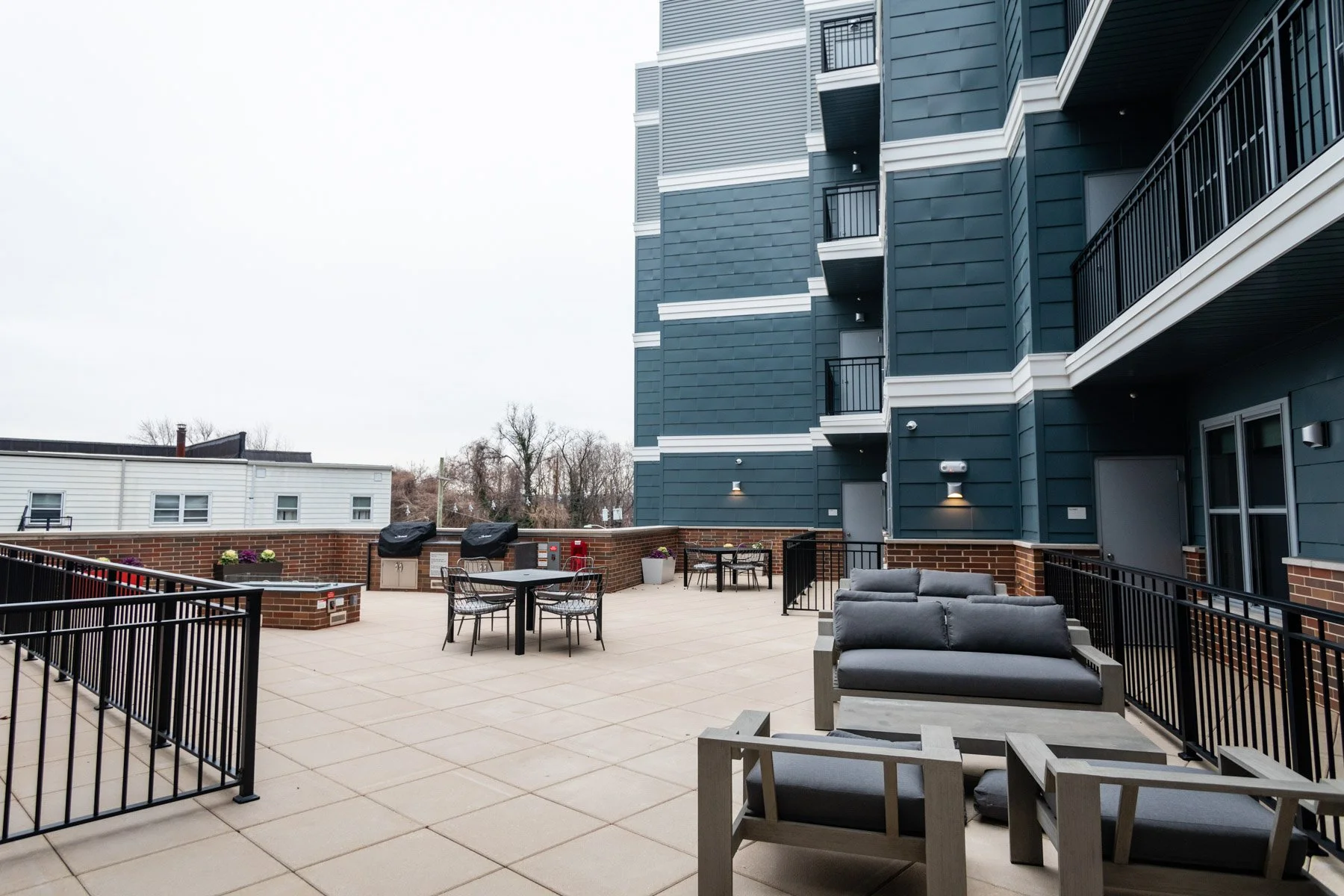 Outdoor apartment balcony with seating and grilling area, featuring dark blue building exterior, brick accents, lounge chairs, a table with chairs, and potted plants, on a cloudy day.
